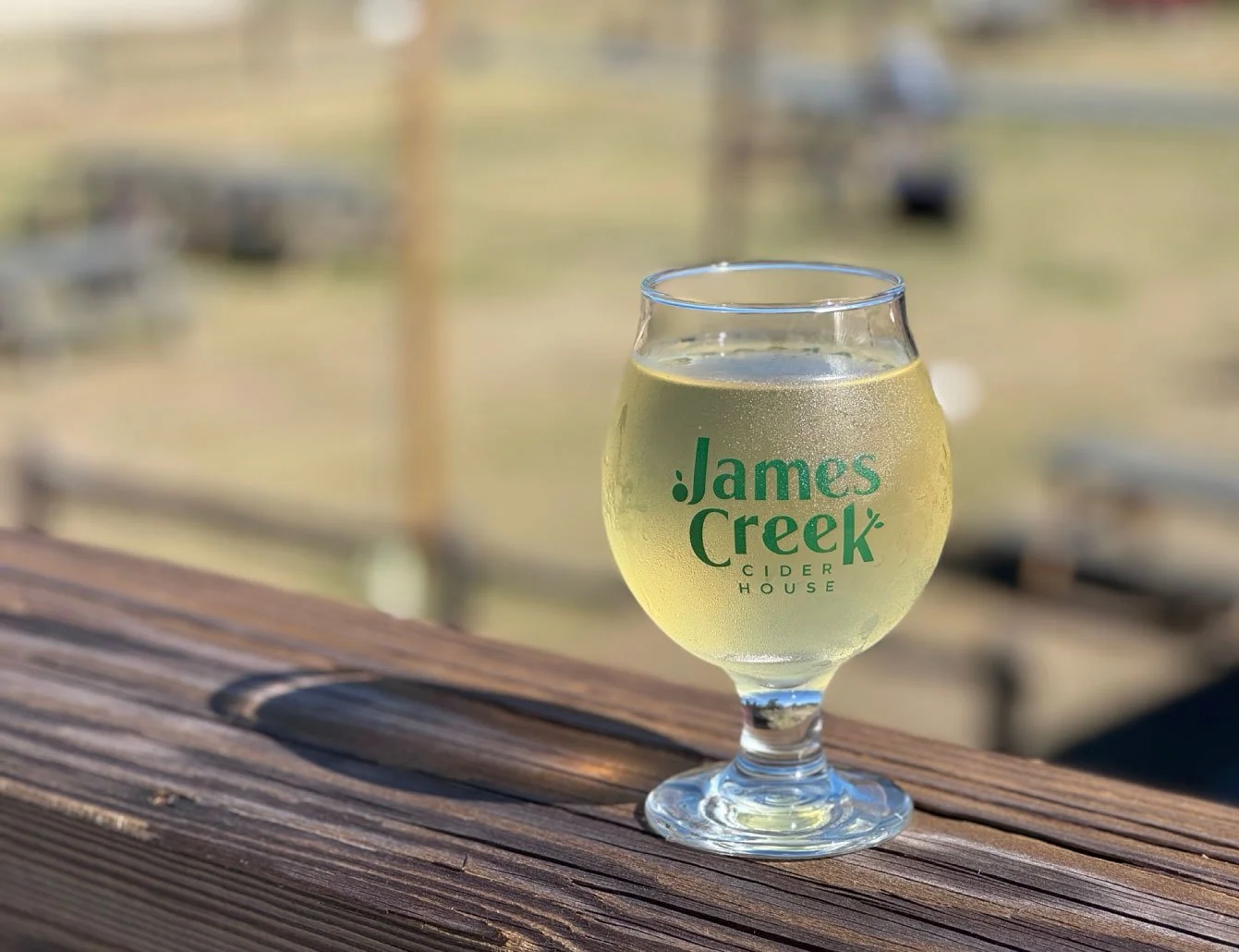 James Creek Cider House branded cider glass — tulip glass with green logo on outdoor wooden railing