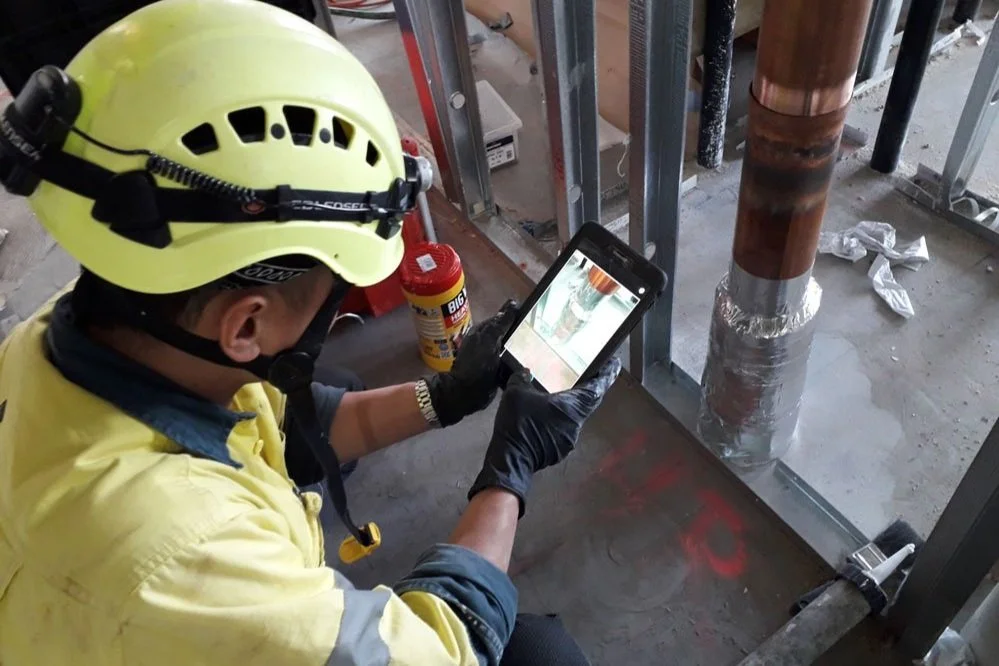 Construction worker in a yellow helmet and jacket taking a photo of a pipe with a smartphone at a construction site.