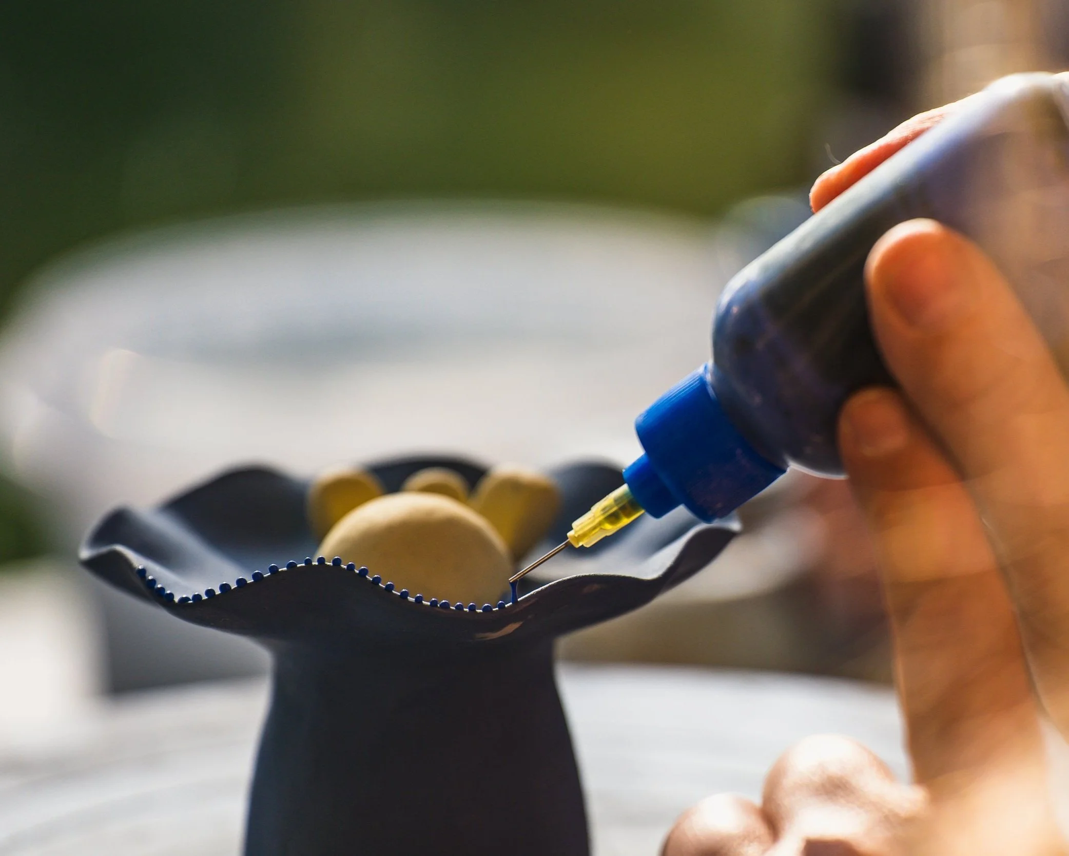 PIcture of an artists hand gently placing a drop of clay sliponto a blue and yellow sculpture