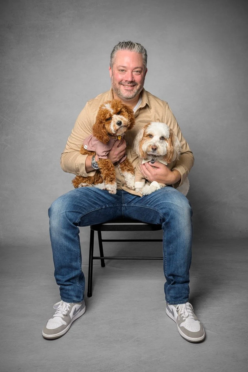A man with short gray hair and a beard, wearing a beige shirt and blue jeans, sitting on a black chair against a gray studio background, holding two dogs, one brown with curly fur and dressed in a pink shirt, and the other white with tan markings, wearing a tan sweater.
