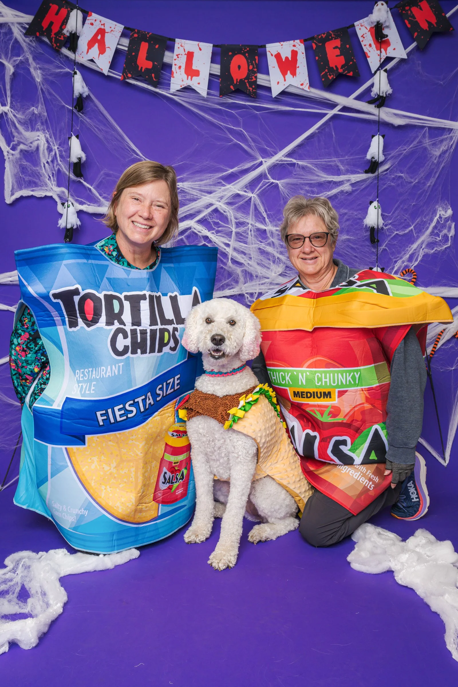Two women and a dog dressed in costumes pose for a Halloween photo in front of purple background decorated with spider webs and hanging ghost and bat decorations, with a Halloween banner overhead.