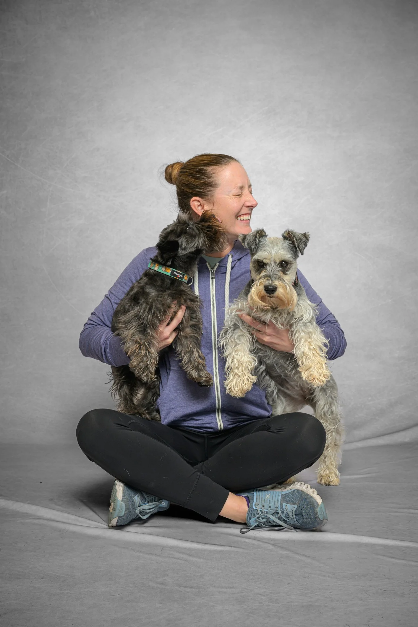 A woman sitting cross-legged on the floor, holding two dogs, smiling and laughing.