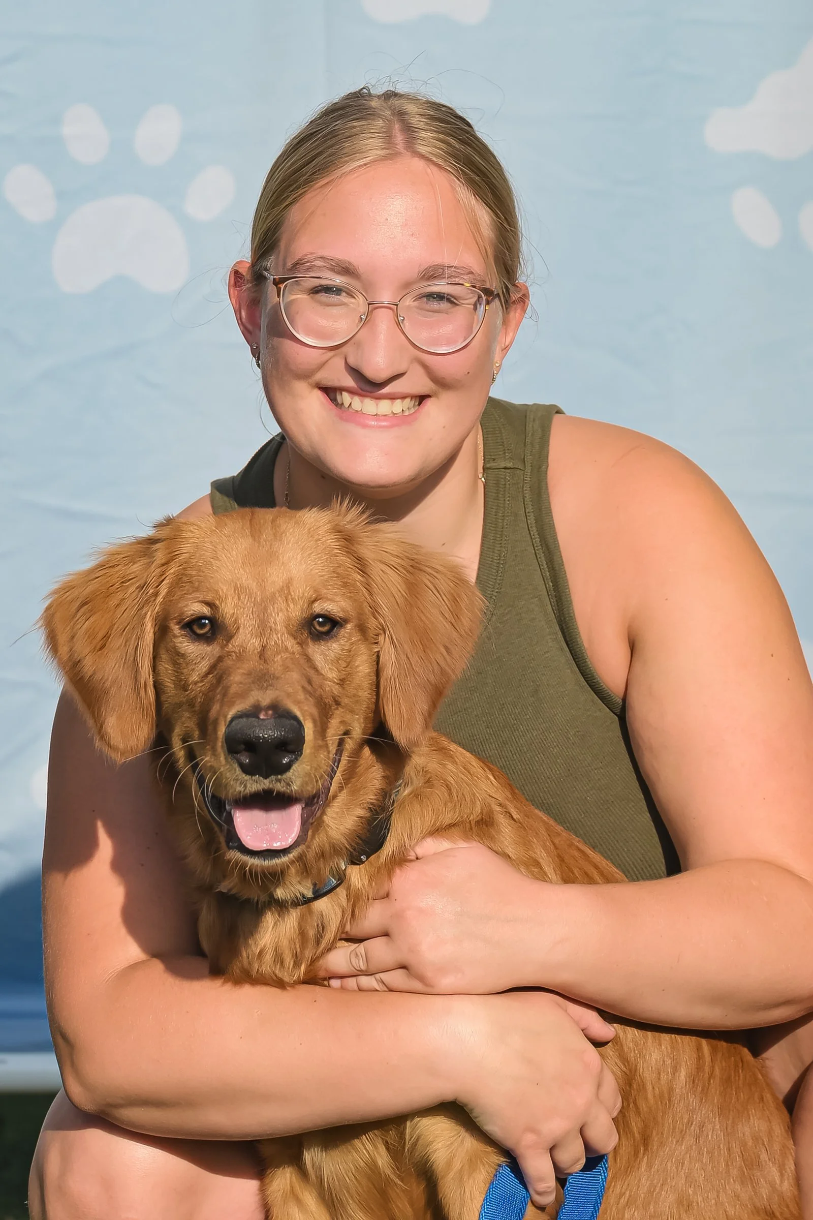 A young woman with glasses smiling, holding a golden retriever. The background features paw prints in white on a light blue backdrop.