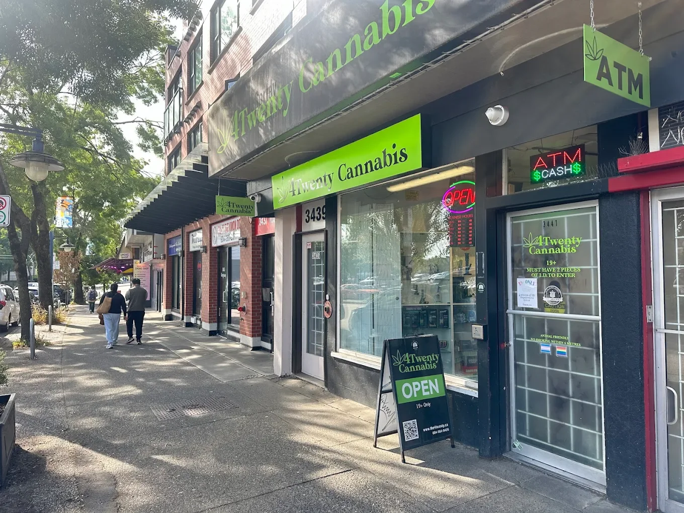 Street view of a storefront called '4Twenty Cannabis' with bright green signage, located at 3441. The store has an 'Open' sign in pink neon and an 'ATM' sign. There is a sidewalk with a few pedestrians walking by, and trees are visible along the street.