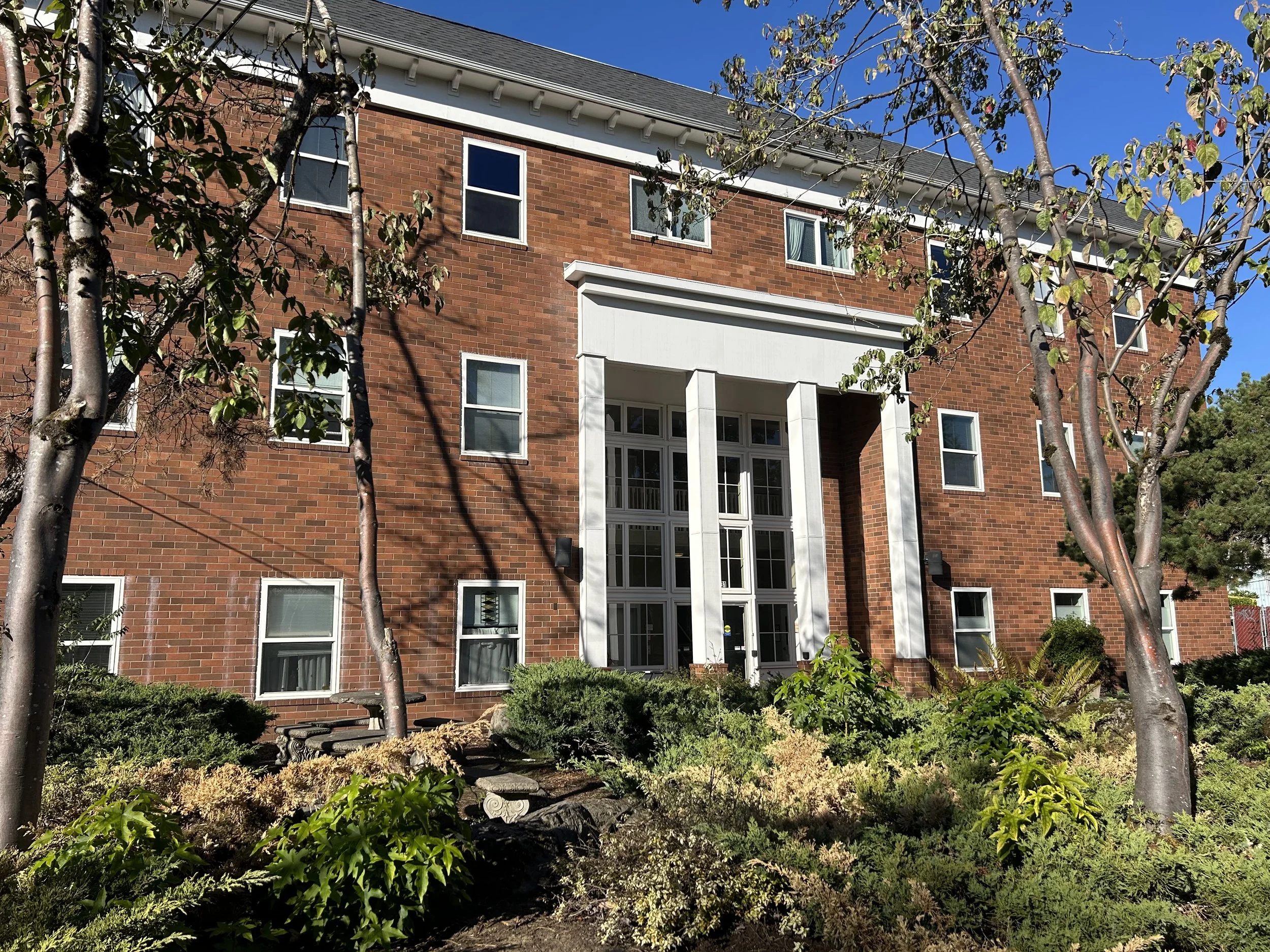 A three-story brick building with white window frames and a central entrance featuring large glass panels and white columns. The foreground includes landscaping with various shrubs and trees.