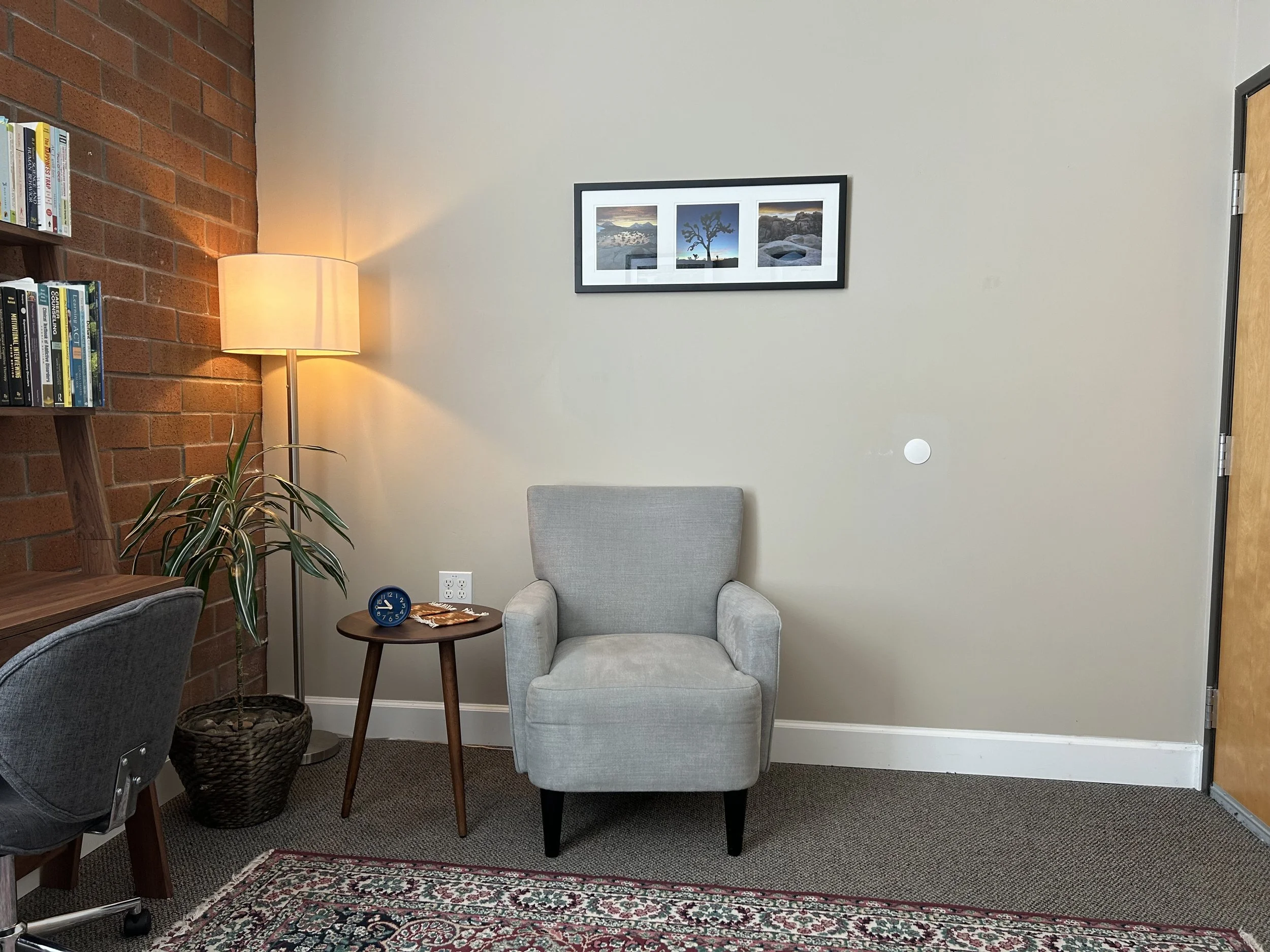 A cozy reading corner with a gray armchair, a small wooden side table with a clock, and a floor lamp. A plant is next to the lamp, and there's a framed photo on the wall. Books are on a shelf against a brick wall, and a rug covers part of the floor.