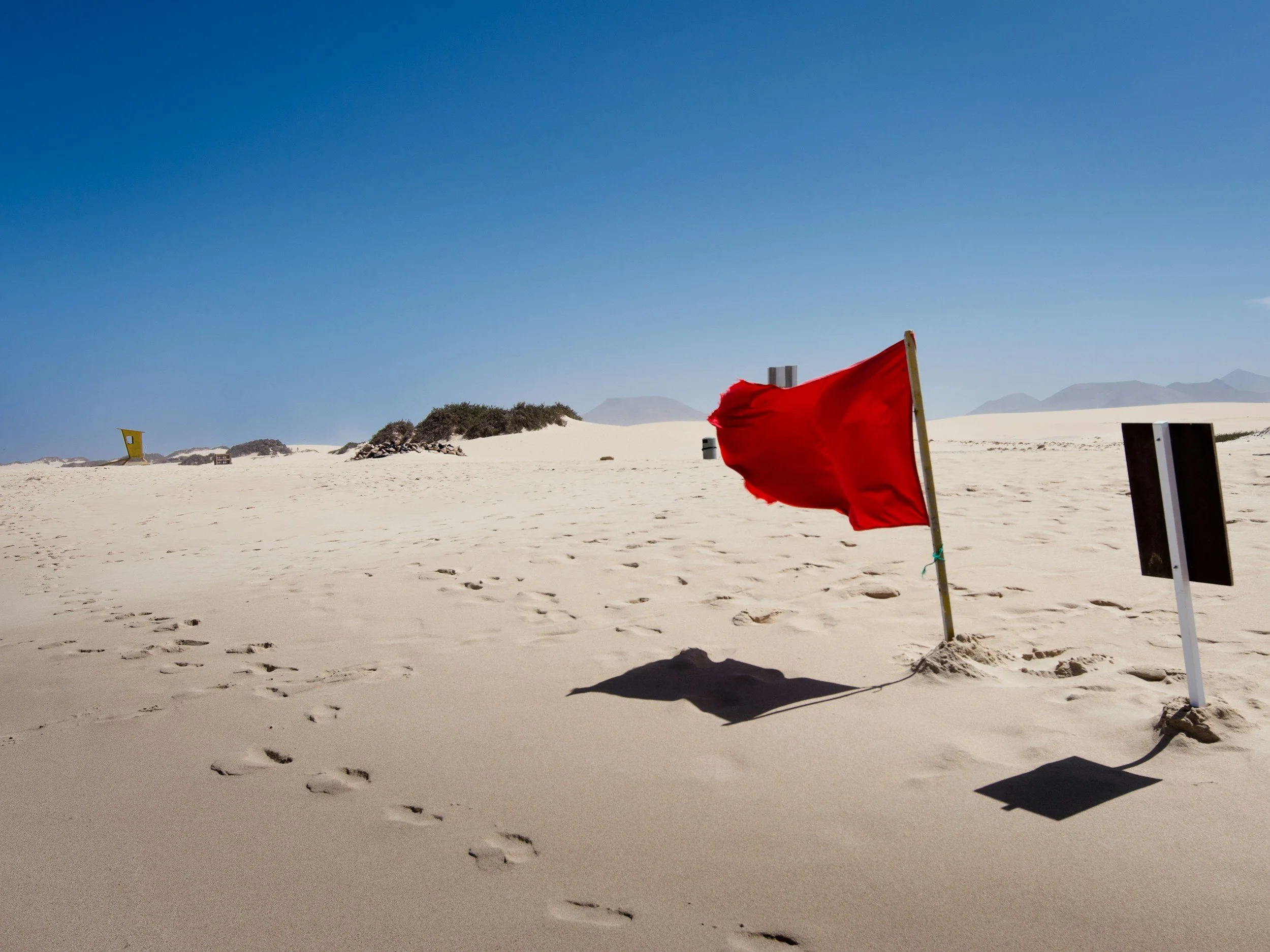 A sandy beach with footprints, a red flag on a pole casting a shadow, and a distant landscape with bushes and mountains under a clear blue sky.