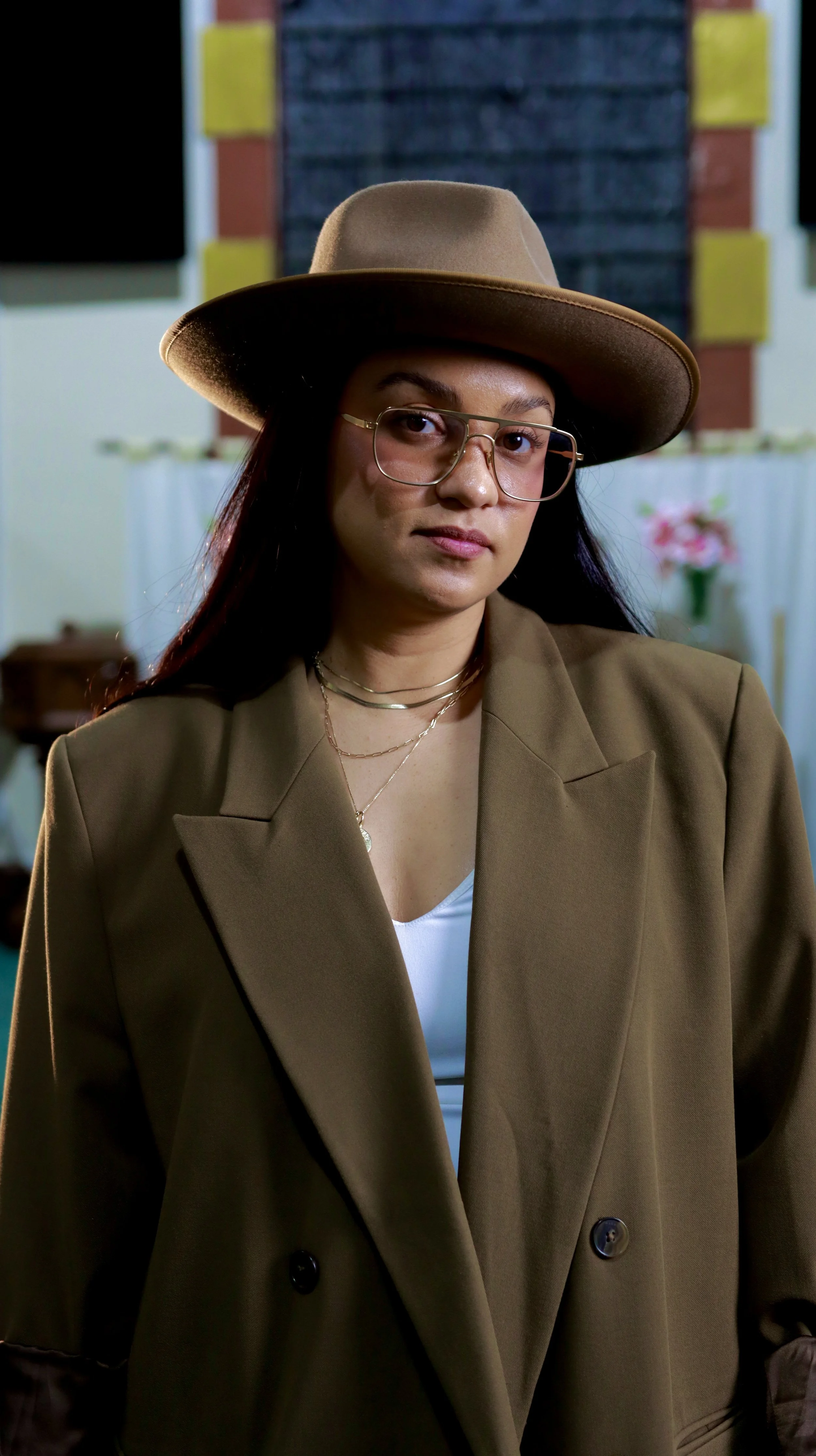 A woman wearing a brown blazer, white shirt, gold-rimmed glasses, layered necklaces, and a tan wide-brimmed hat, standing indoors with blurred background.