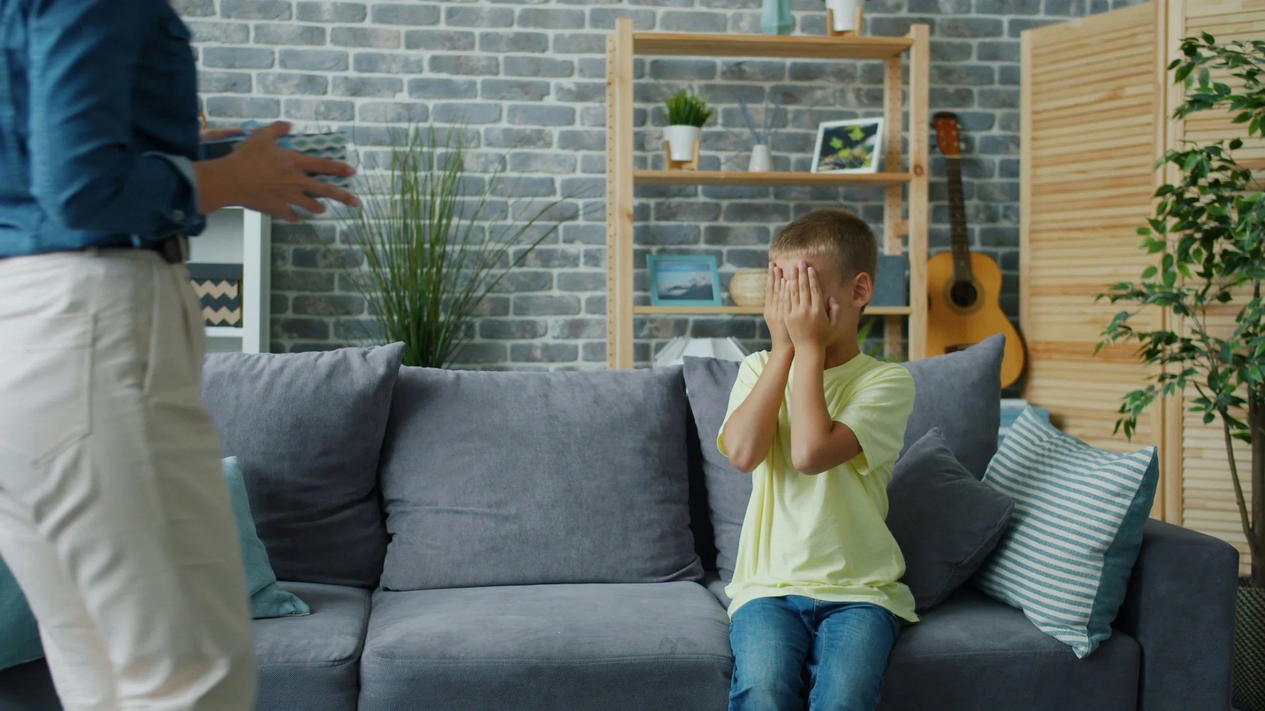 Young boy sitting on a couch covering his face while an adult stands nearby in a living room