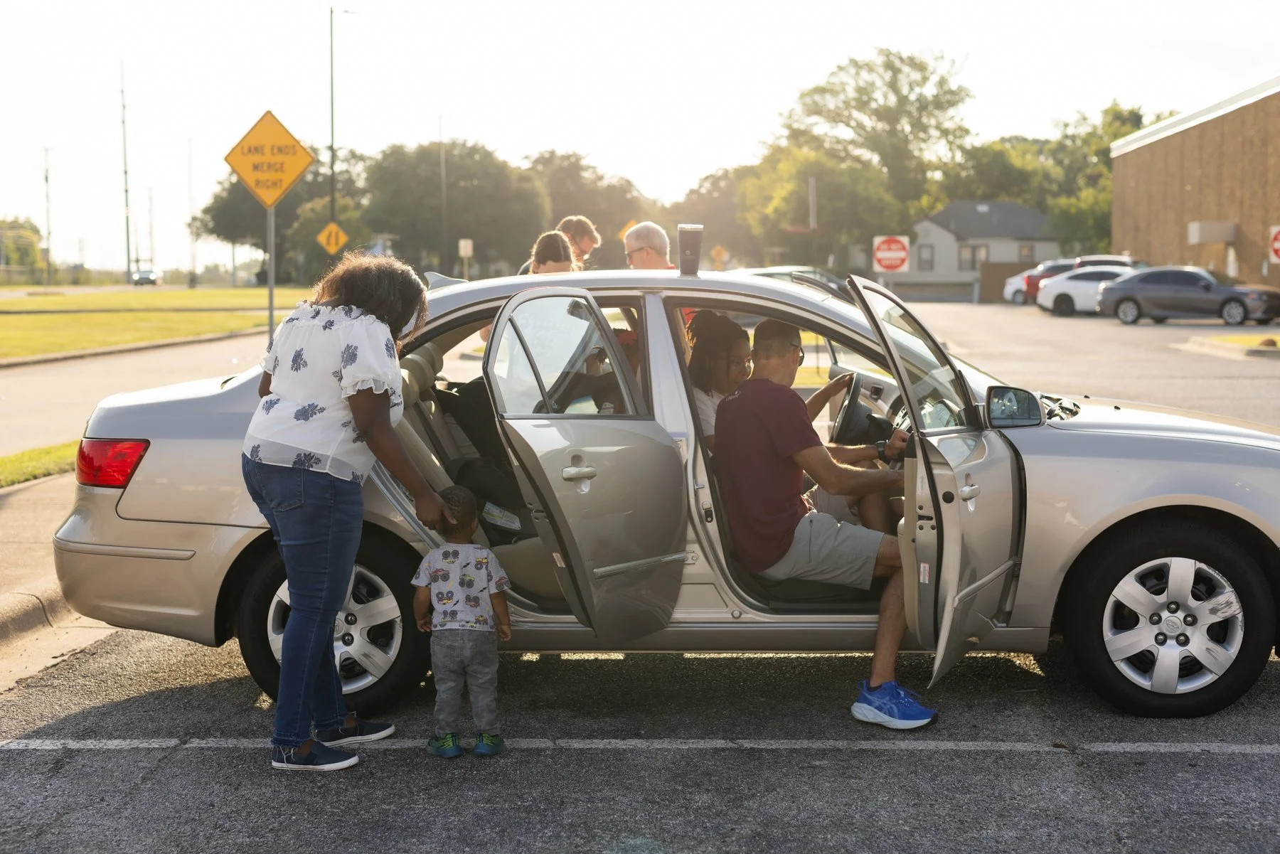 Family gathering around a donated silver car in a parking lot during daylight, with a single mom and two children near the car and others inside.