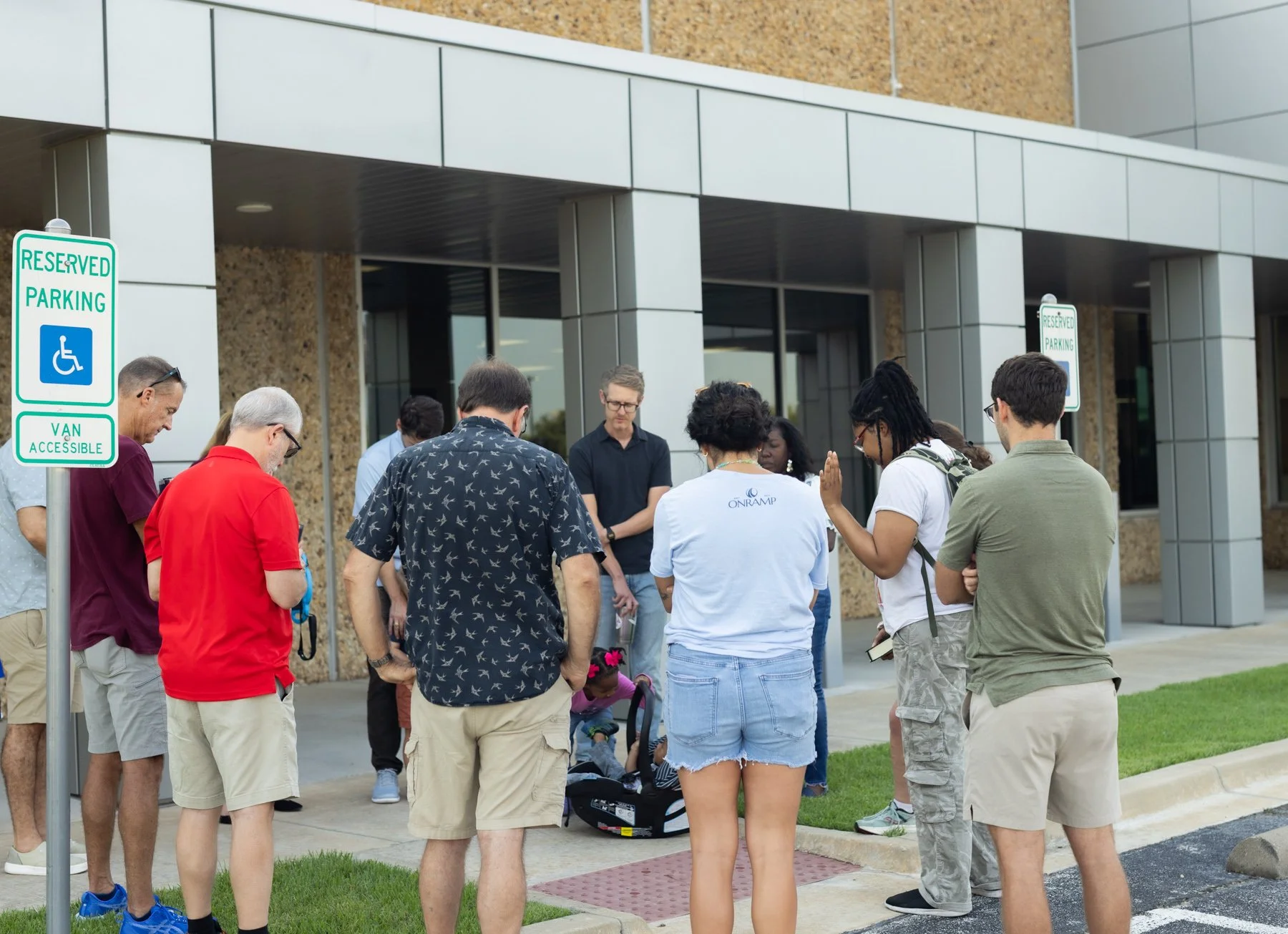A group of people gathered outside a building, and praying in thankfulness for a newly donated car.