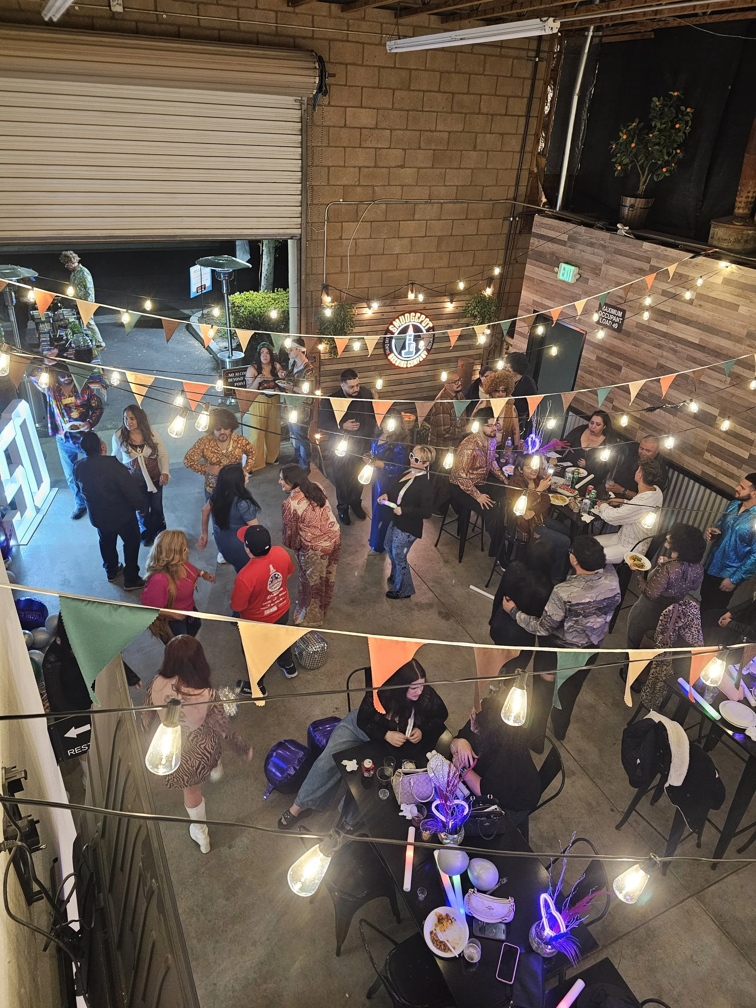 People gathering at a social event or party inside a venue with string lights, bunting flags, and a wooden wall with a neon sign that says 'SMOKEBOAT'. Some individuals are dining, chatting, or standing around, with a view into the outdoor area through a partially open garage-style door.
