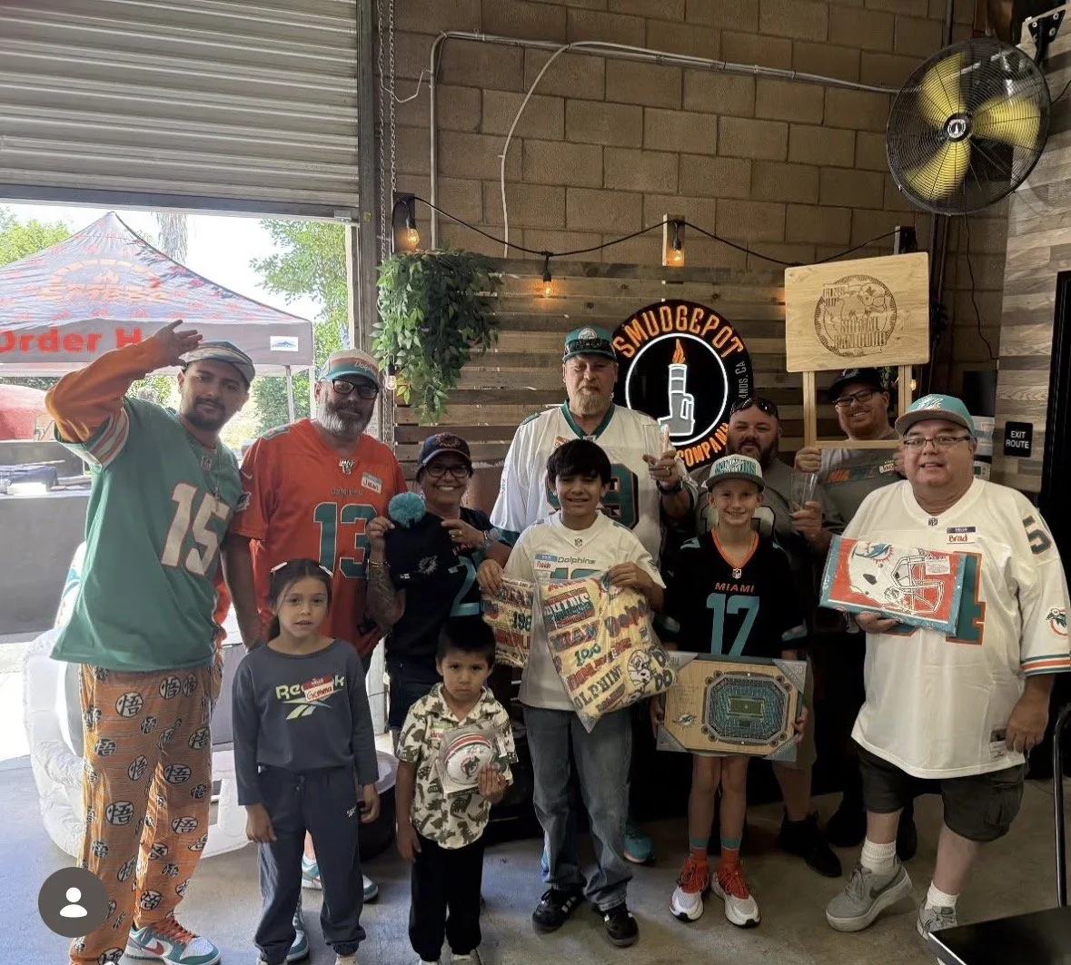 Group of people in a bar or restaurant, some wearing Miami Dolphins jerseys and hats, gathered for a celebration or event with football-themed items and decorations.