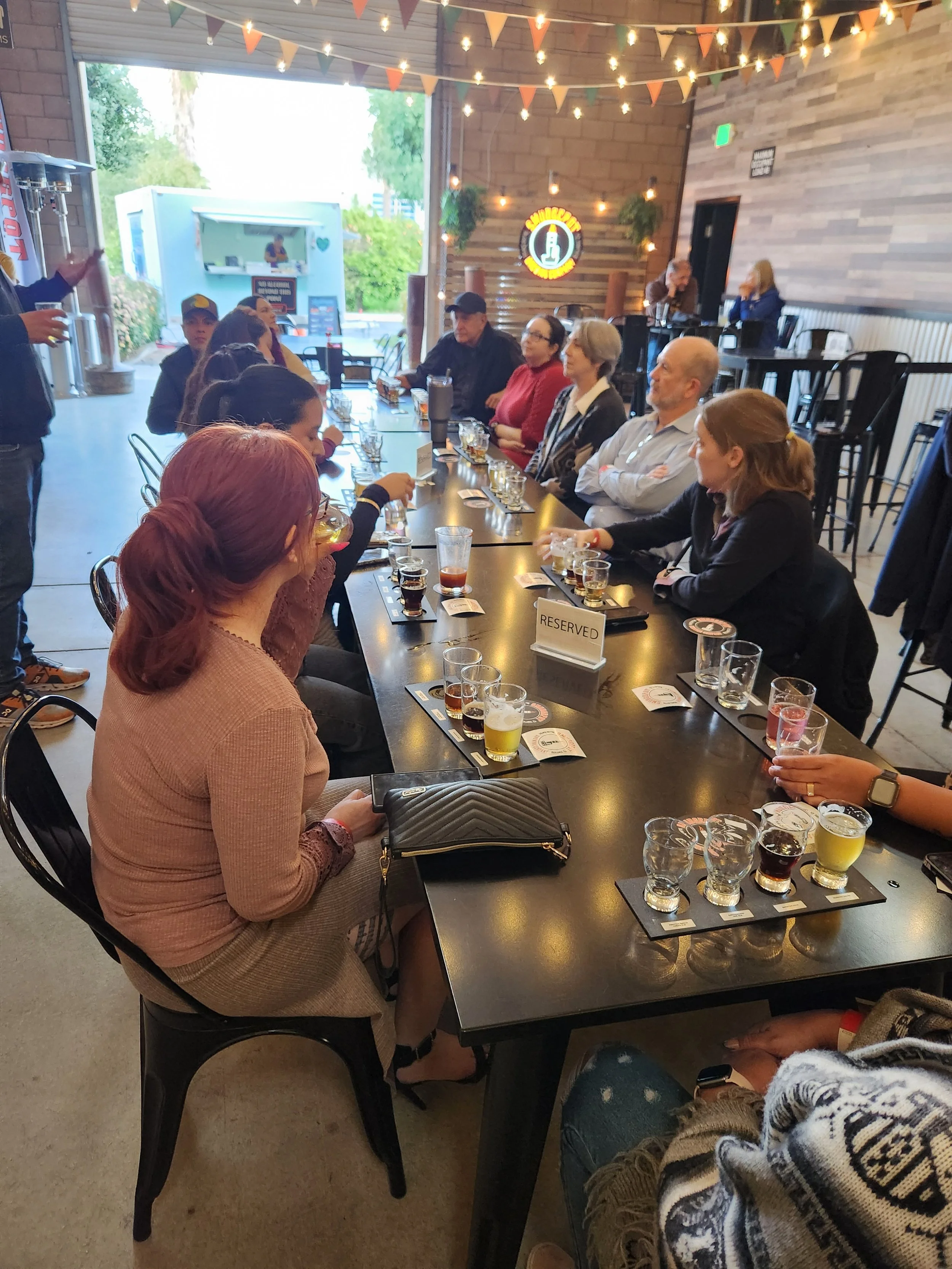 People participating in a beer tasting event at a brewery with multiple small glasses of beer on a long table, a host explaining, and a reserved sign on the table.
