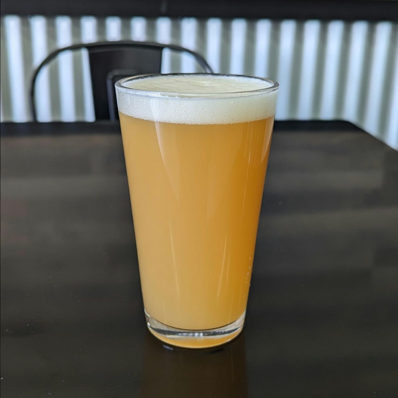 A tall glass of hazy, light yellow beer with a frothy white head, placed on a dark table with a blurred background of black chairs and a balcony railing.
