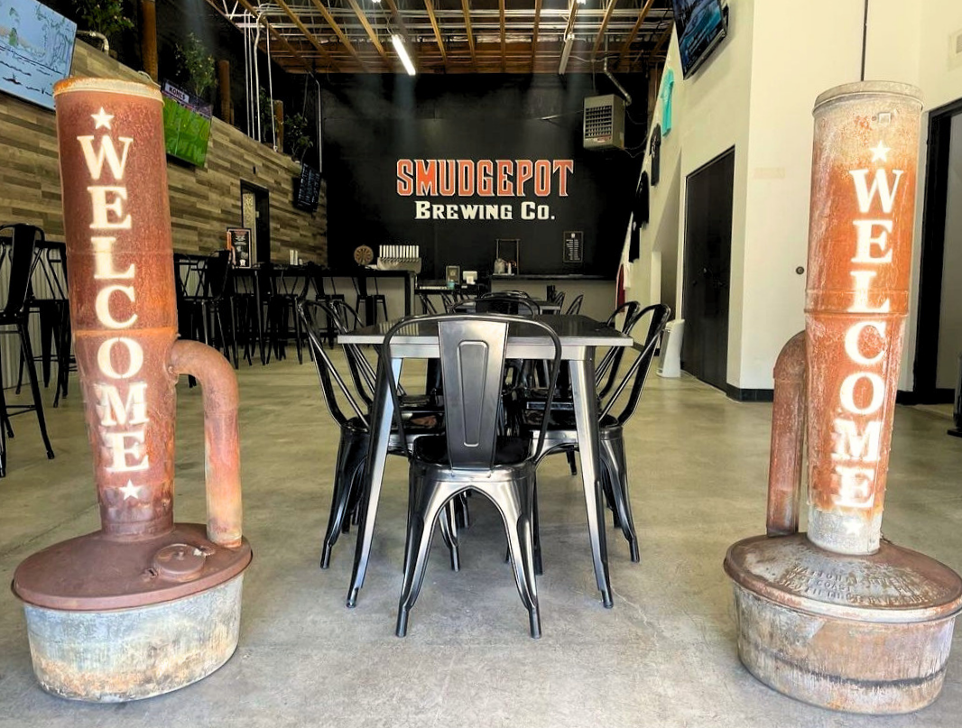Indoor view of a brewery named Smudgepot Brewing Co., with two large rusty welcome beer taps, a round black table with black chairs, a black wall with the brewery's name, bar area in the background, and flat-screen TVs on the walls.