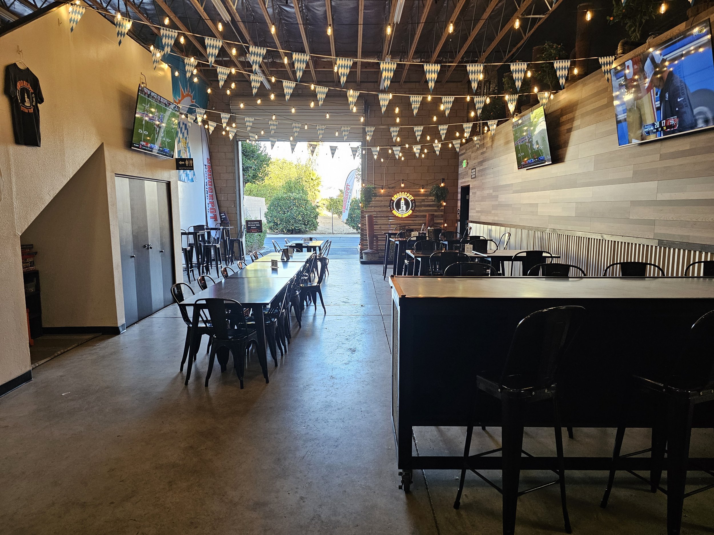 Empty seating area inside a restaurant with a high ceiling, dim lighting, string lights, and a large open door showing outdoors with trees and a parking lot. Multiple flat-screen TVs mounted on the walls display sports games, and a sign for SauDeSport is illuminated on the back wall.