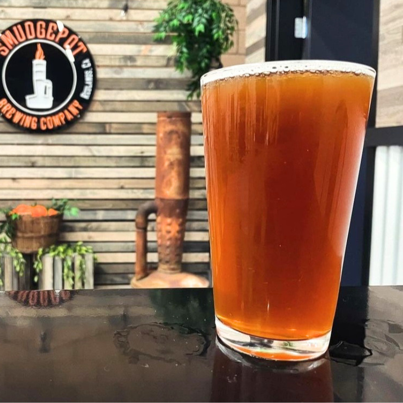 A pint glass filled with amber-colored beer on a dark table in front of a patio with wooden siding, a potted plant, and a chimney with a logo for Smuggler Brewing Company.