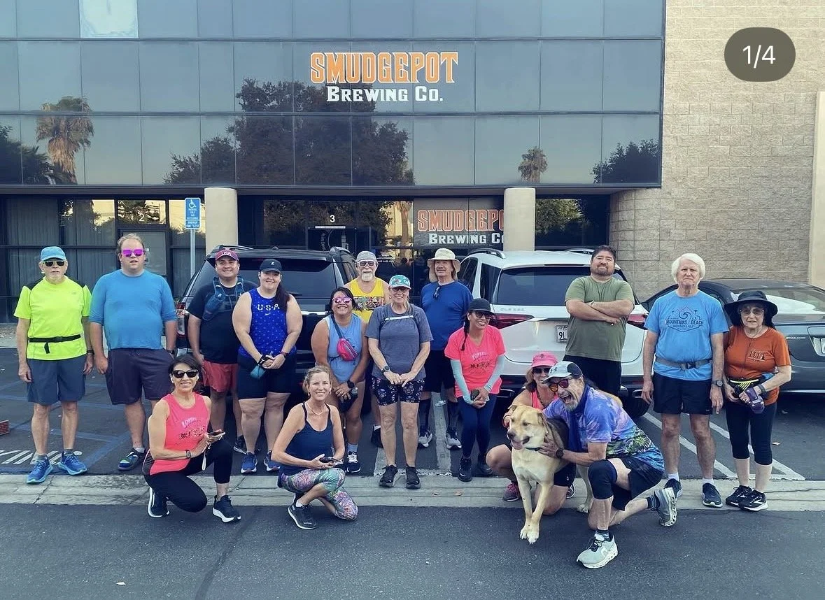 Group of people standing and kneeling in front of the Smudgepot Brewing Co. building, posing for a photo, some with sunglasses and athletic gear, with a dog in front.