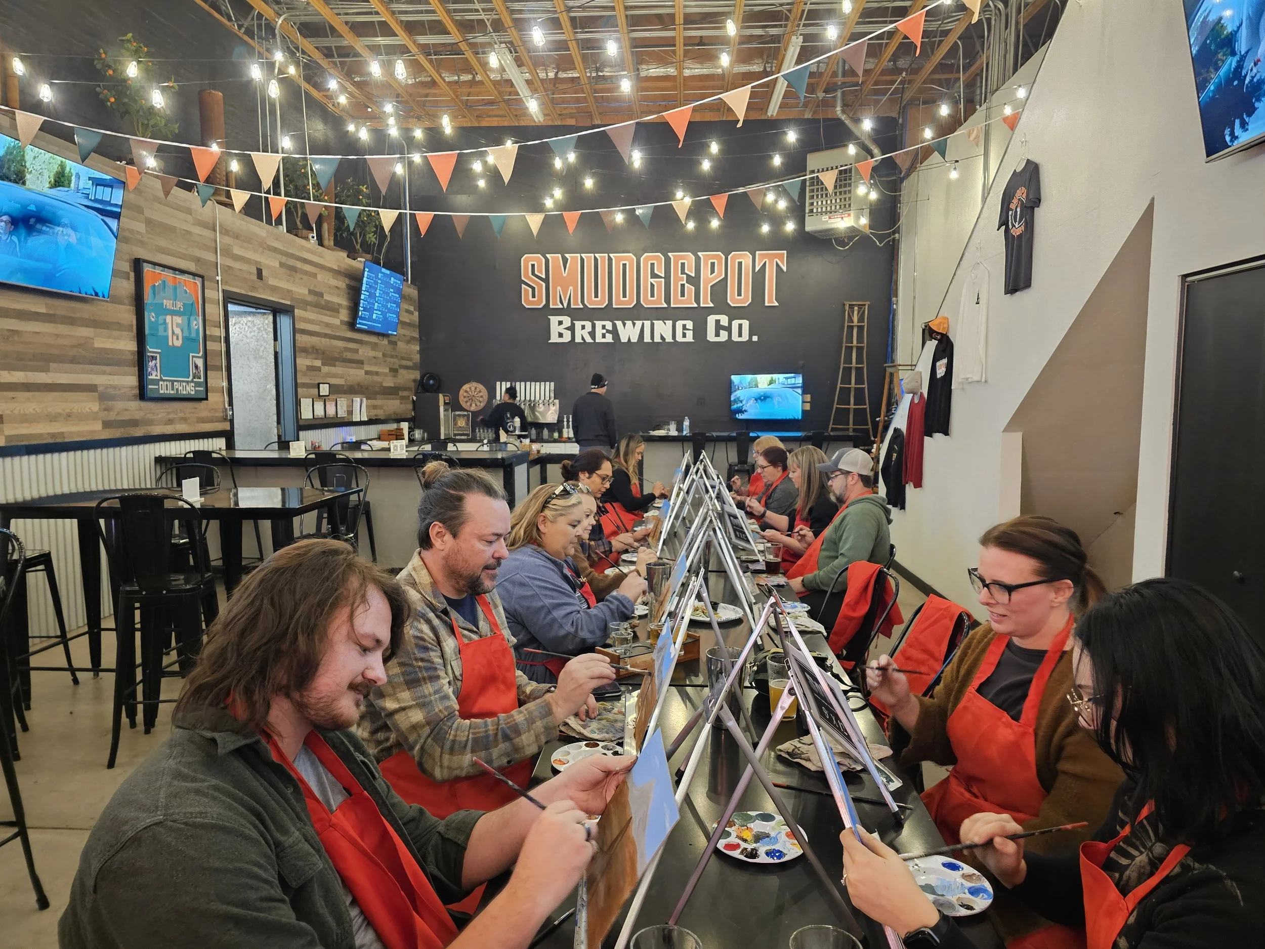People seated at art tables painting at Smudgepot Brewing Co., decorated with string lights and colorful bunting, with a wooden wall and television screens in the background.