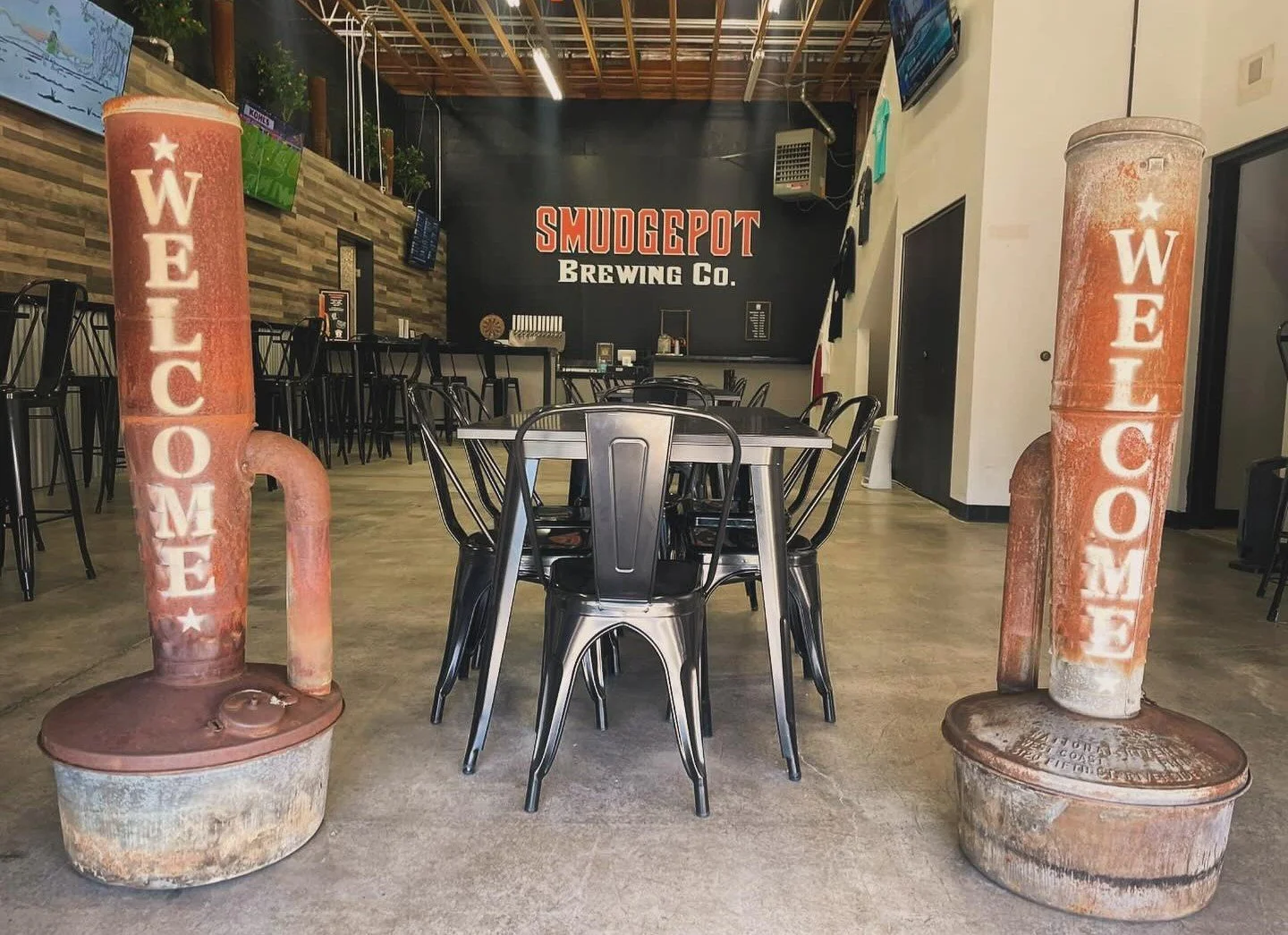 Interior of a brewery with tables and chairs, featuring two large fake pipe signs with 'WELCOME' written on them, at Smudgepot Brewing Co.