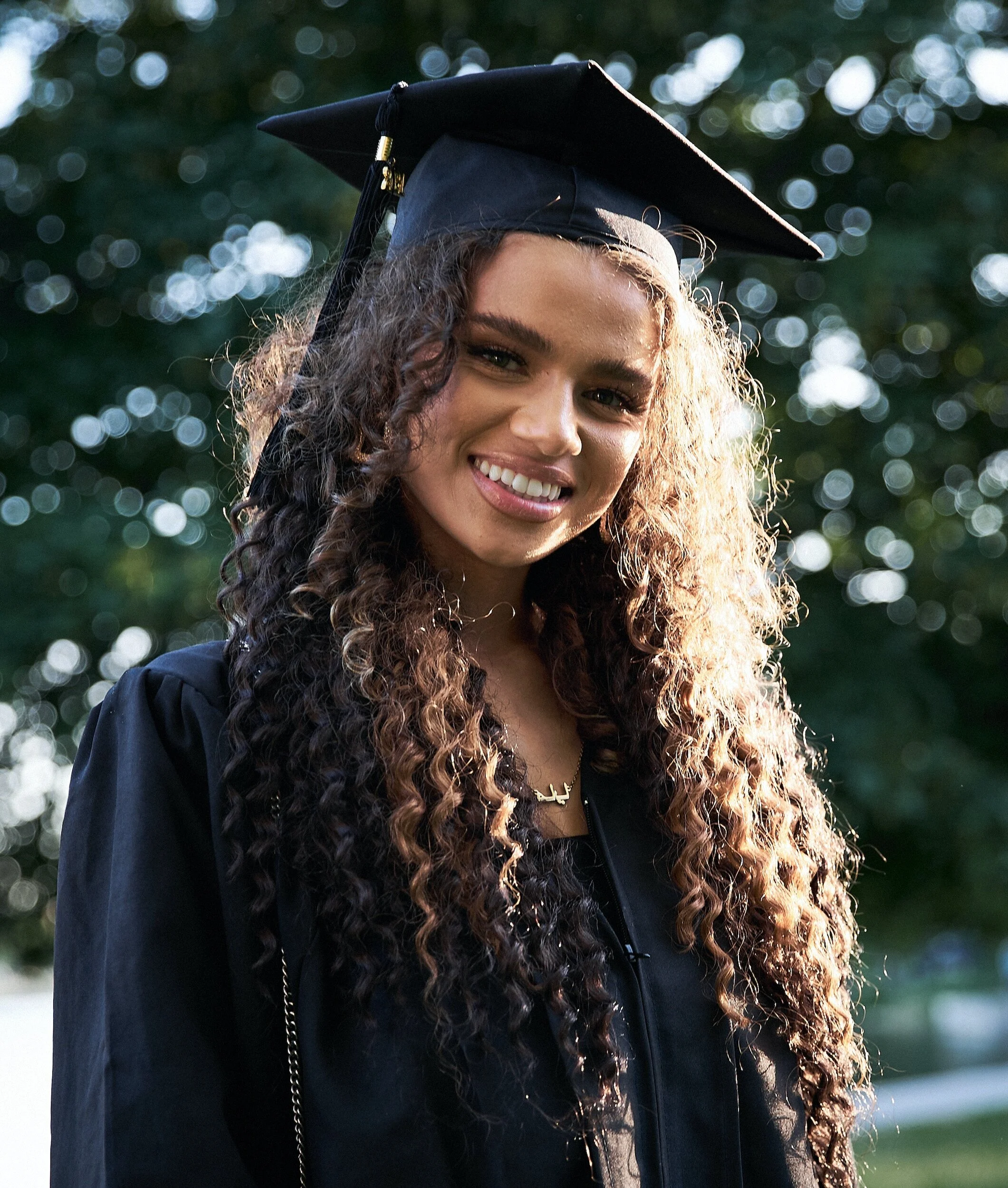 A young woman in a graduation cap and gown smiling outdoors with trees and blurred lights in the background.
