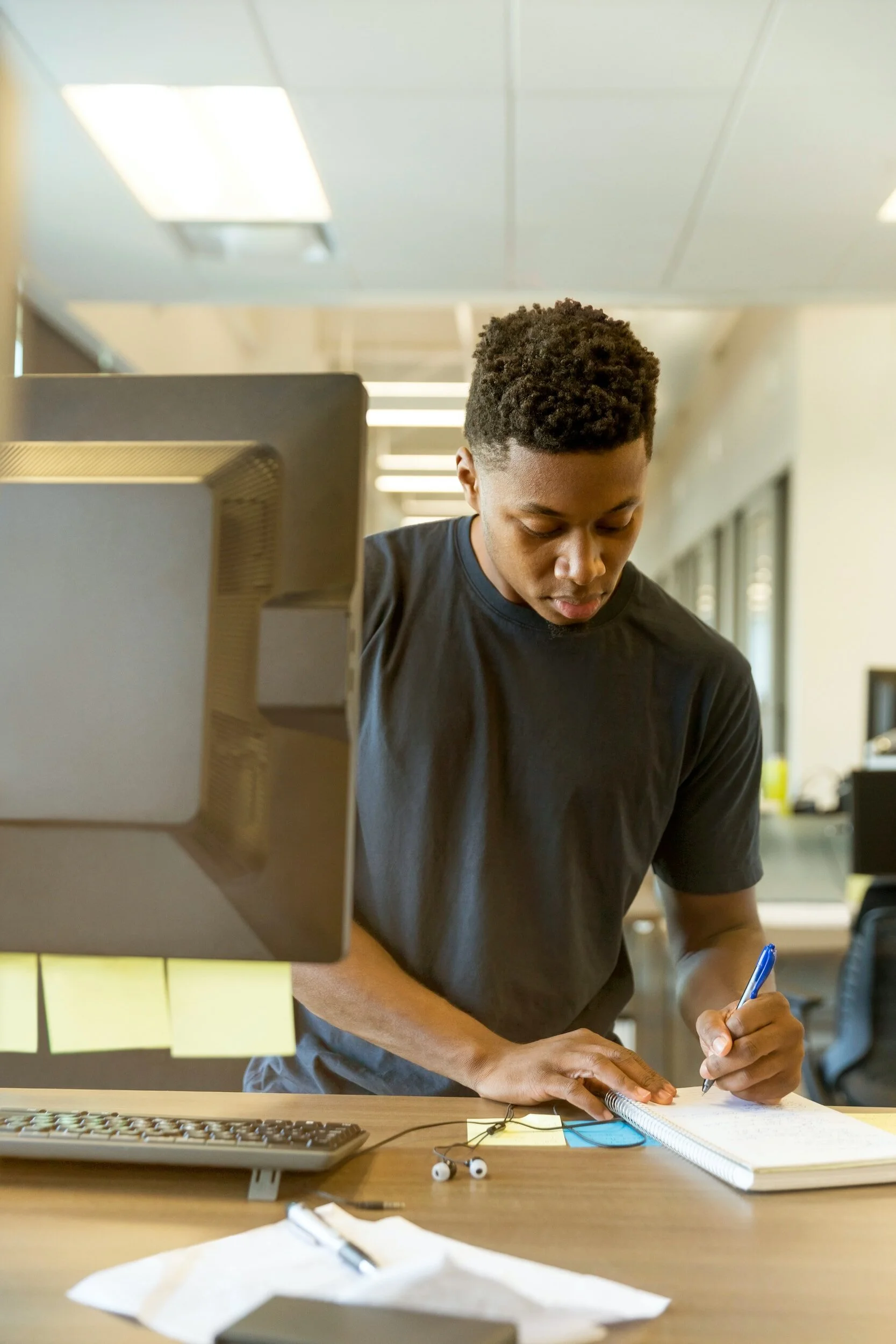 Young man in a black t-shirt writing in a notebook at a cluttered desk in an office.