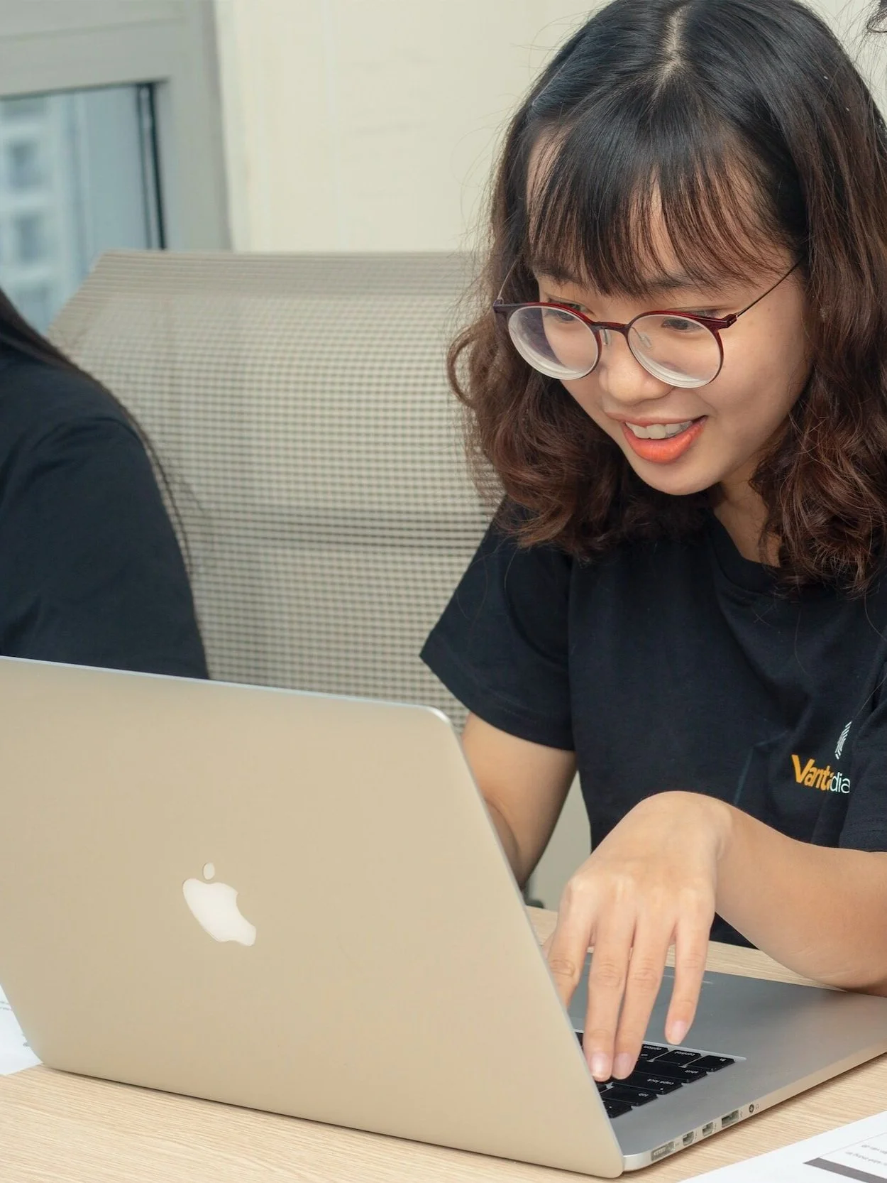 A young woman with wavy brown hair, glasses, and wearing a black shirt, working on a silver MacBook laptop at a desk, smiling as she interacts with the computer.