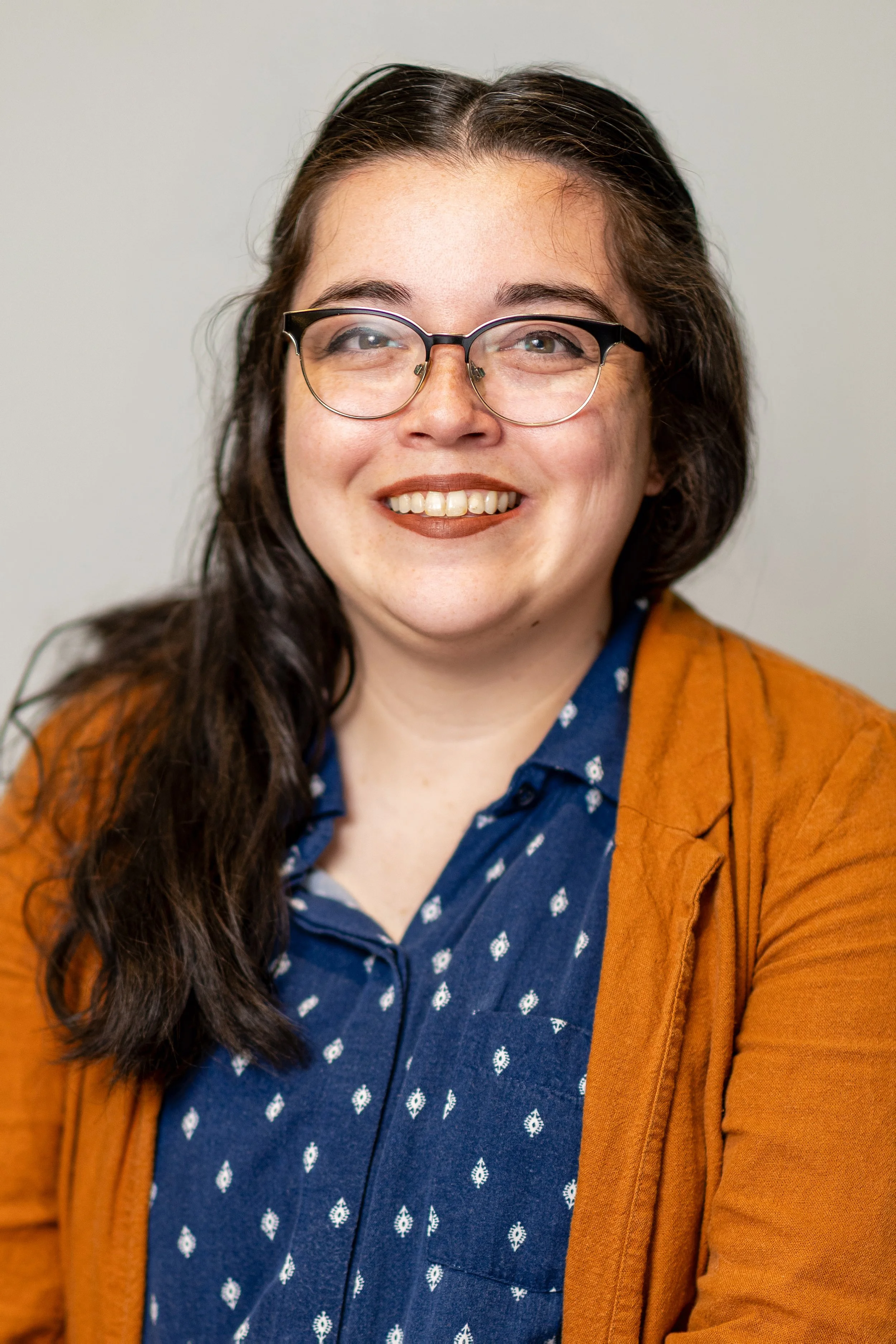 Portrait of a woman with shoulder-length dark hair, glasses, and earrings, smiling against a green background.