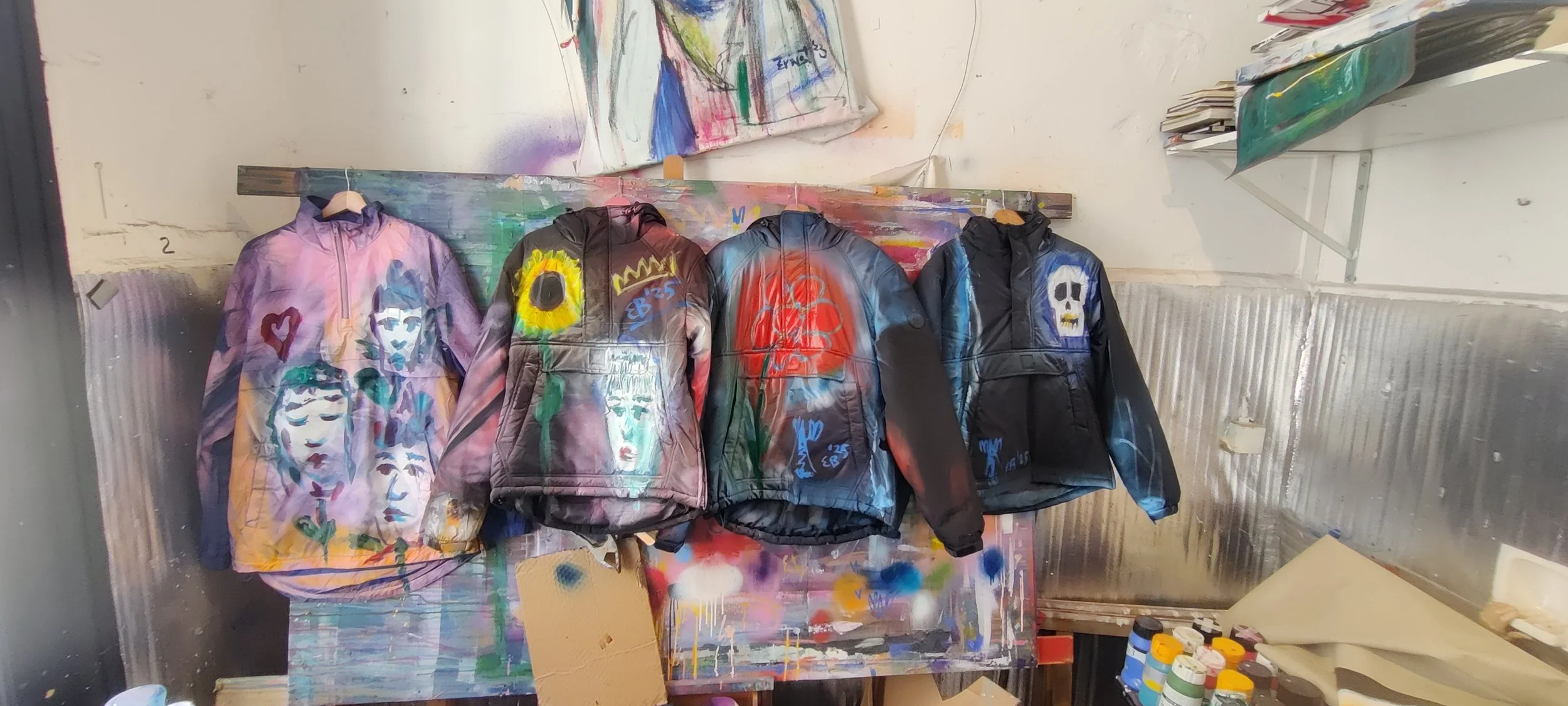 Four jackets hanging on a rack, each with graffiti-style artwork including faces, flowers, skulls, and symbols, inside an art studio with paint supplies and artwork in the background.