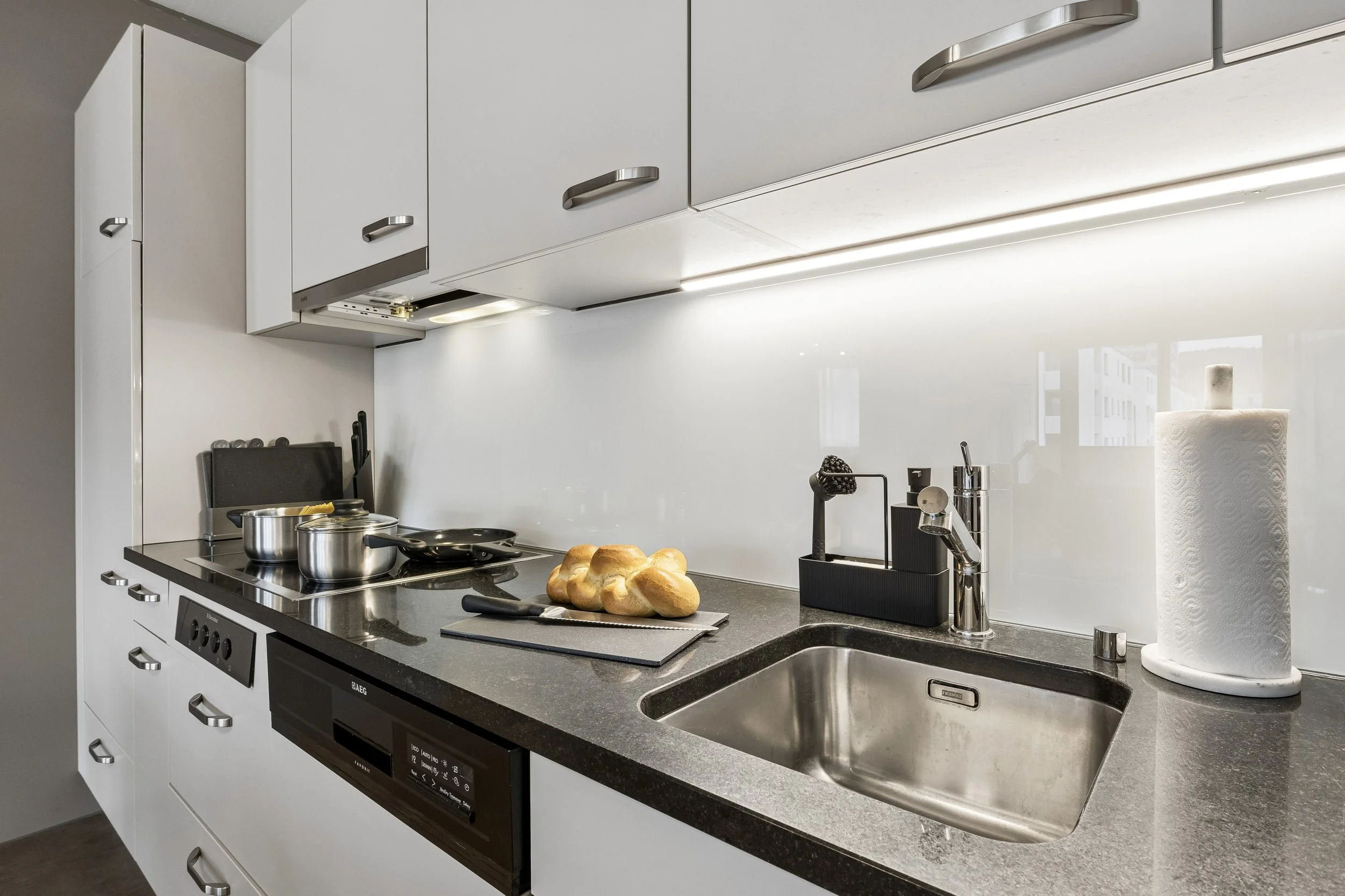 A modern kitchen with white cabinets, a black countertop, a stainless steel sink with a paper towel roll nearby, a stove with pots, bread rolls on a cutting board, and various kitchen utensils.