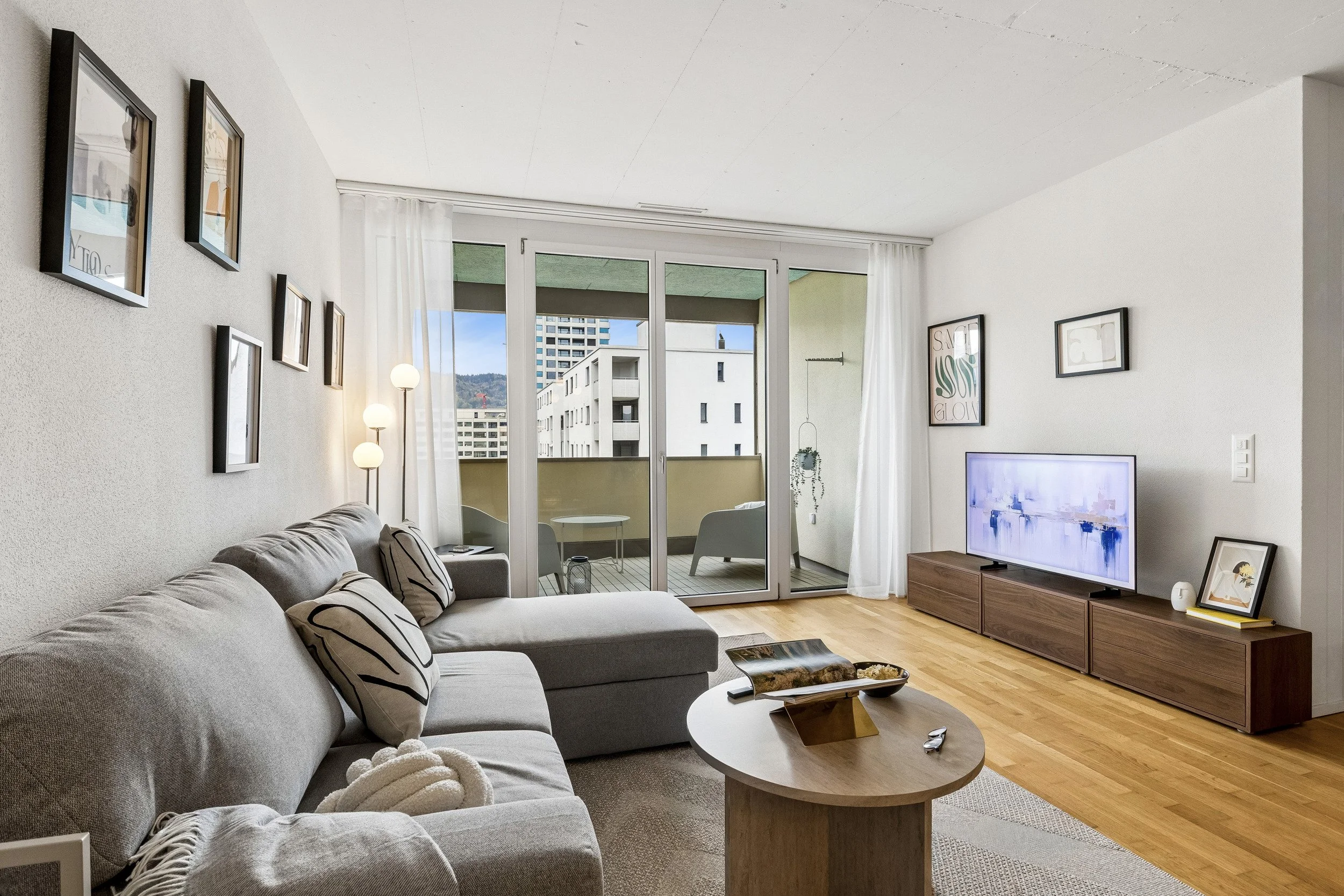 Living room with a gray sofa, paintings on the wall, a wooden TV stand with a TV, a round coffee table, and large sliding glass doors opening to a balcony with outdoor seating.