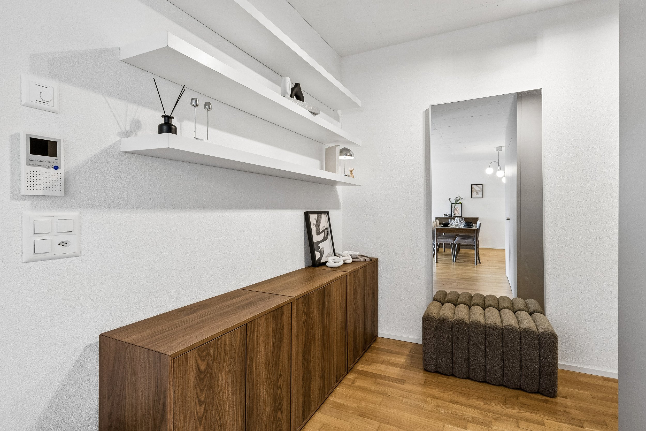 A hallway with a wooden sideboard, white floating shelves with decorative items, and a small upholstered bench. A doorway reveals a dining area with a table and chairs, hanging pendant lights, and wall art.