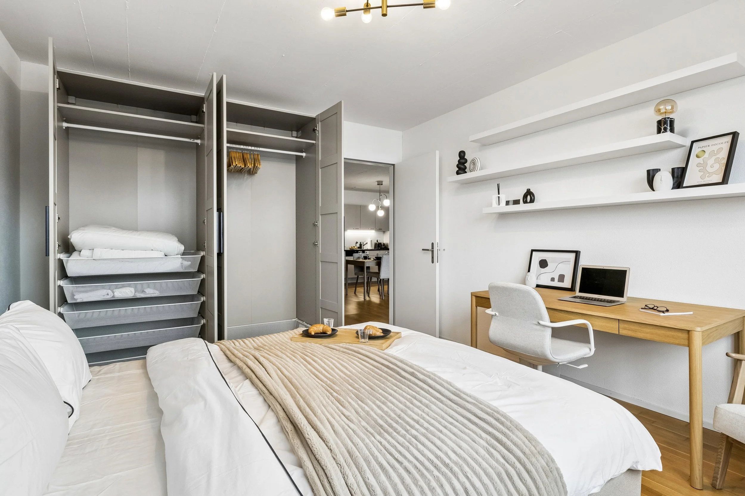 Modern bedroom with open closet, white bed with beige blanket and pillows, wooden desk with laptop and glasses, white shelves with decorative objects, and doorway to a kitchen area.