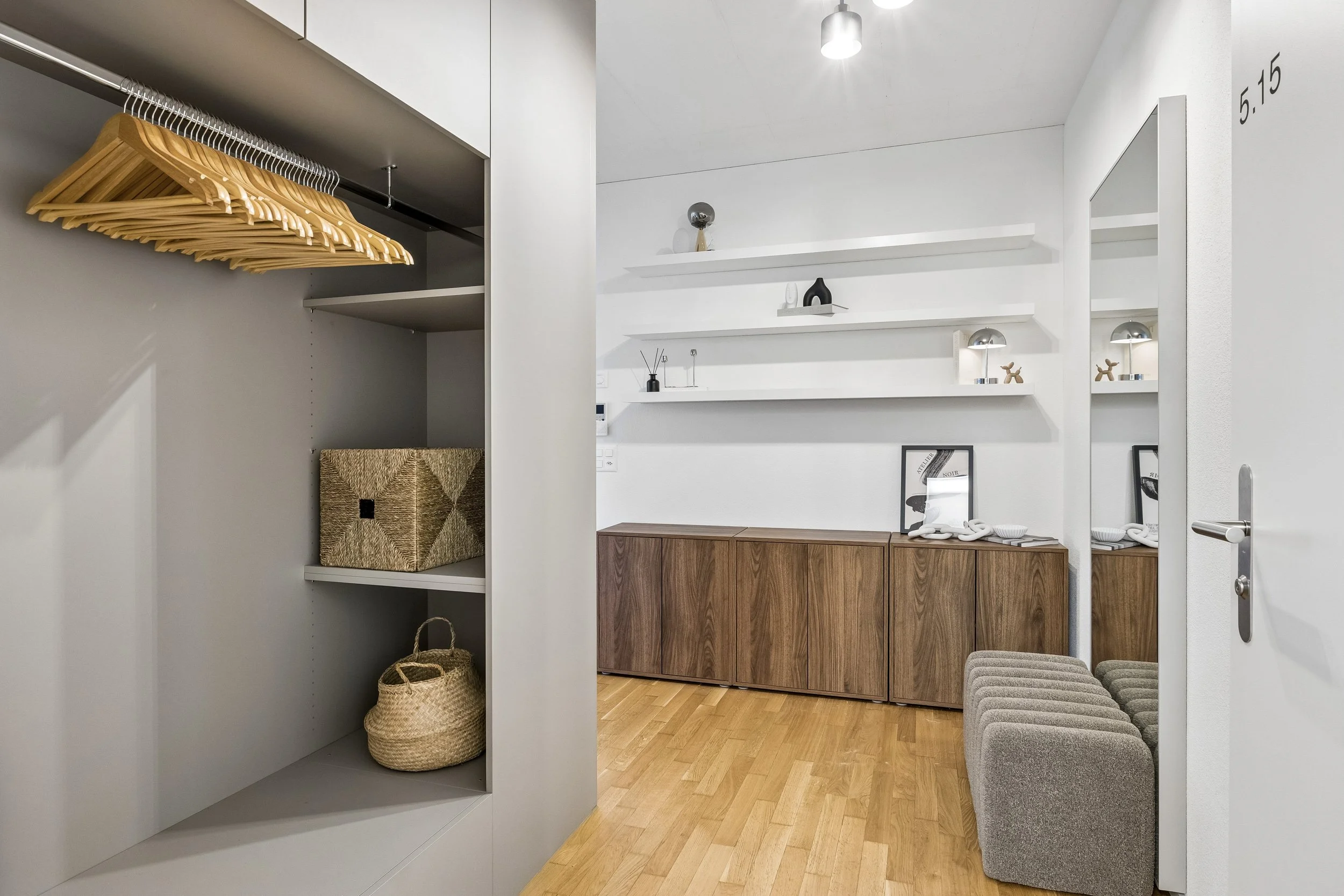 A modern, minimalist closet and dressing area with white walls, floating shelves, wooden cabinets, and beige upholstered stools. There are empty wooden clothes hangers on a metal rail, a woven basket, and a collection of decorative items on the shelv