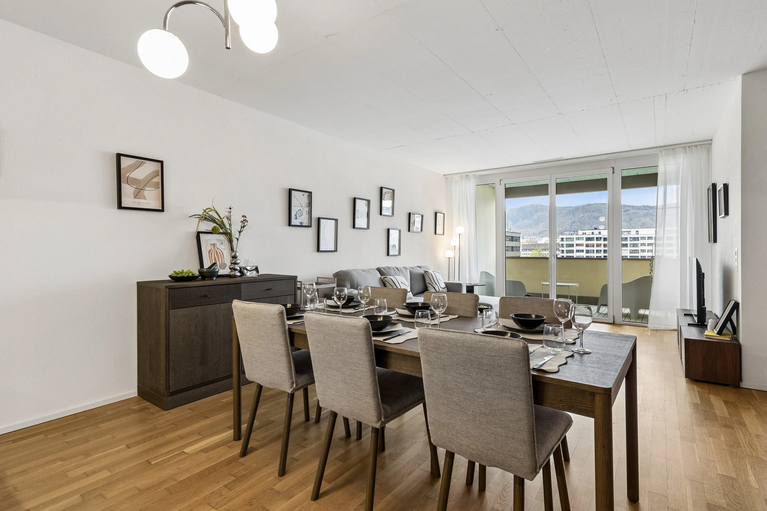 A modern living room with a dining area, featuring a wooden dining table set with black bowls, glasses, and cutlery. Beige upholstered chairs surround the table. The room has a white wall decorated with framed artwork and a dark wooden sideboard with