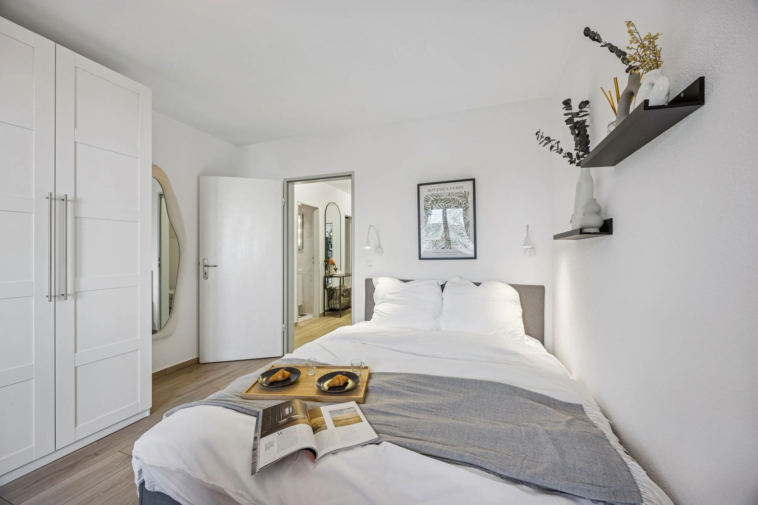 A modern bedroom with a white bed, a bedside table with breakfast, and a wall shelf with vases and decor. An open door reveals part of a bathroom.