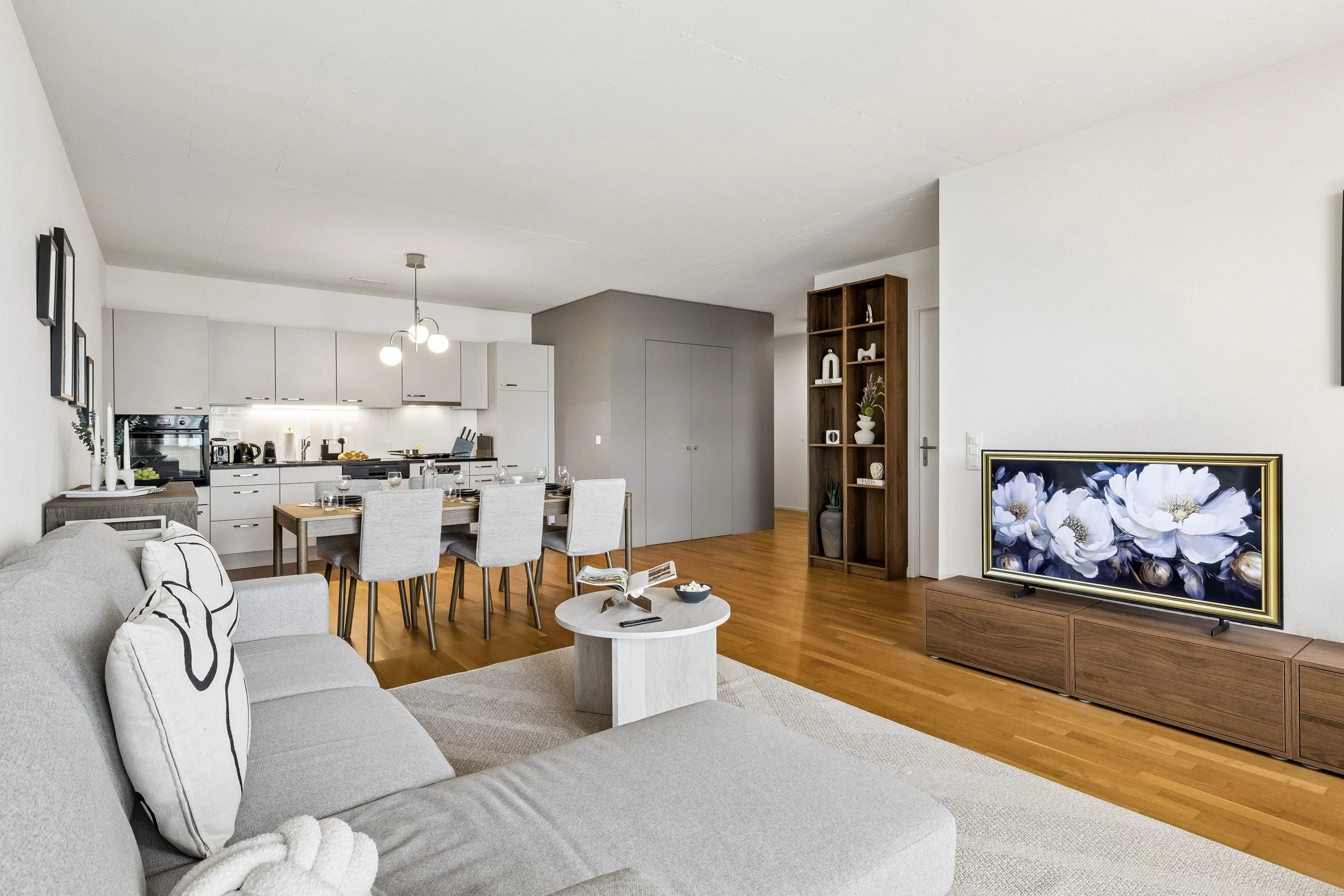 Interior of a modern living and dining room with a gray sofa, wooden coffee table, dining table with chairs, a TV on a wooden stand, and a bookshelf. The kitchen with white cabinets, black appliances, and a dining area are visible in the background.