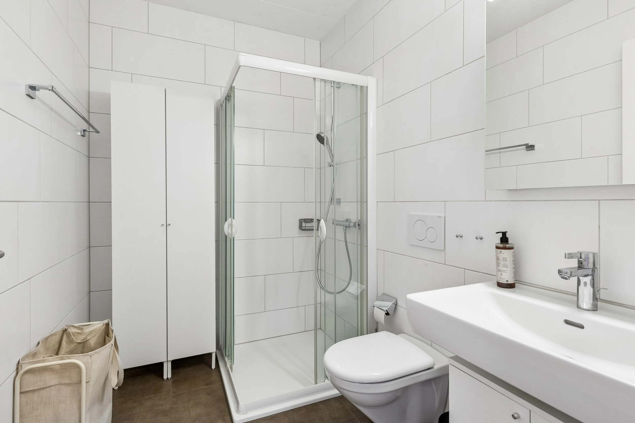 Modern white-tiled bathroom with a shower stall, toilet, small sink, mirror, and beige laundry basket.