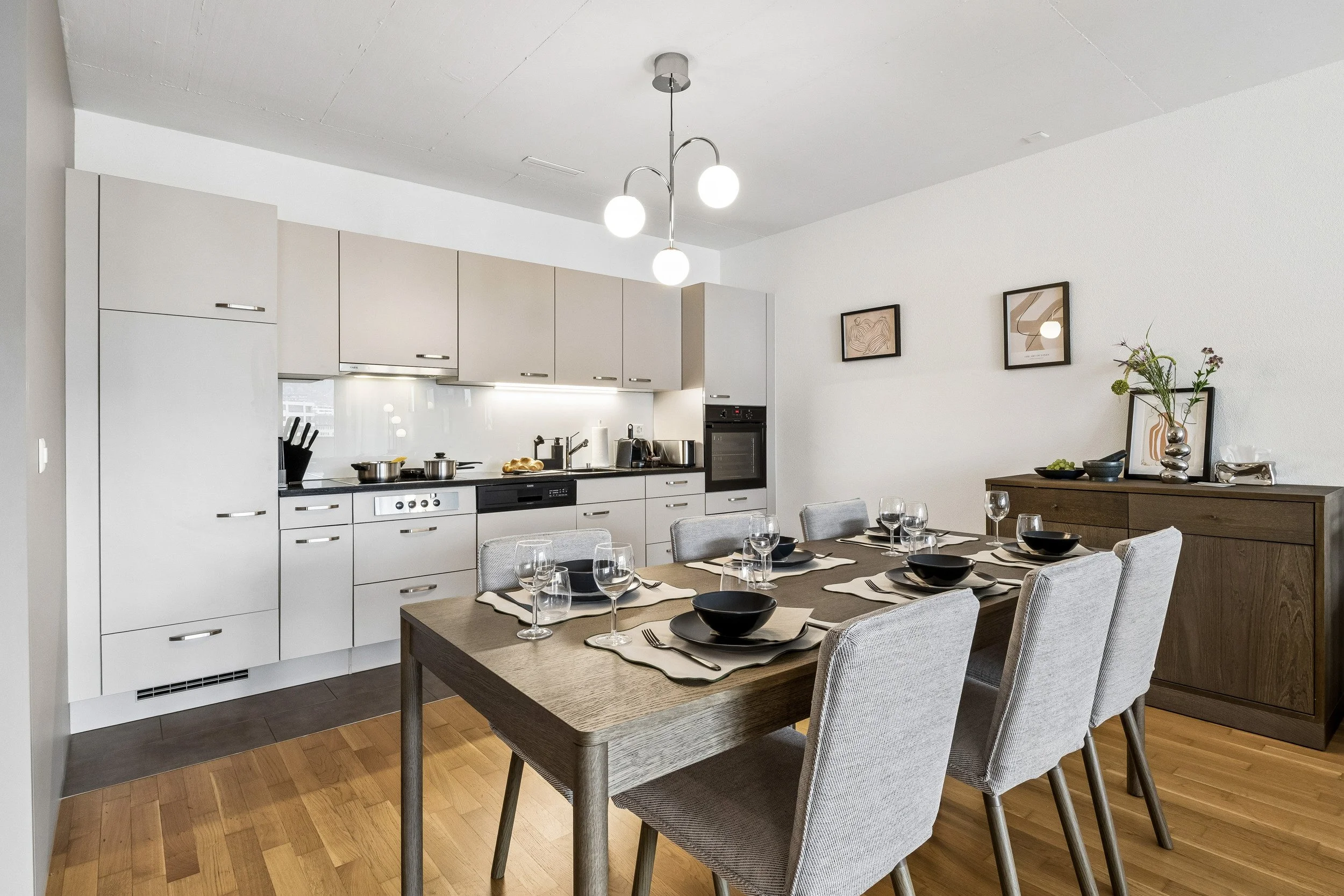Modern kitchen and dining area with a wooden table set with black bowls, wine glasses, and silverware, beige chairs, white cabinets, and minimalist wall art.