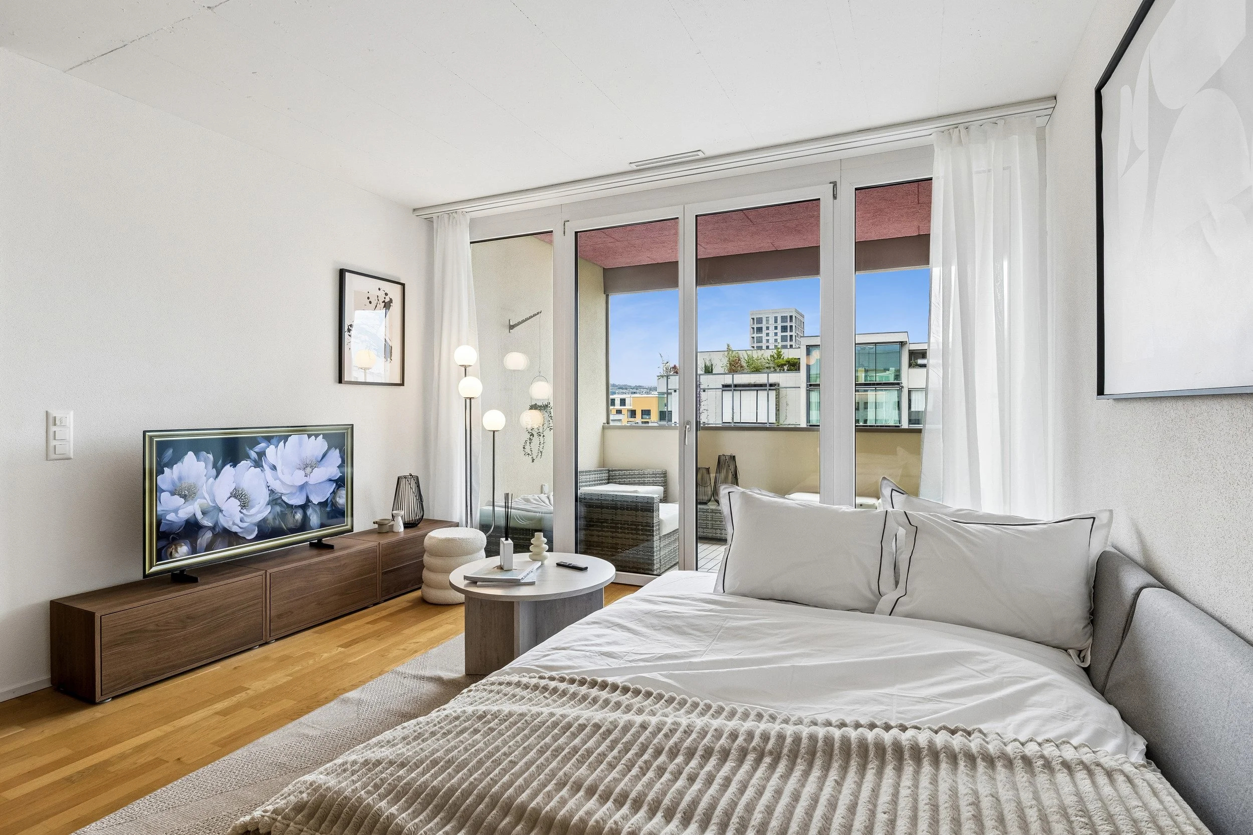 Modern living room with sliding glass doors leading to balcony, white walls, wooden floors, sofa with white pillows, flat-screen TV on wooden stand, abstract artwork on wall, and urban cityscape view.