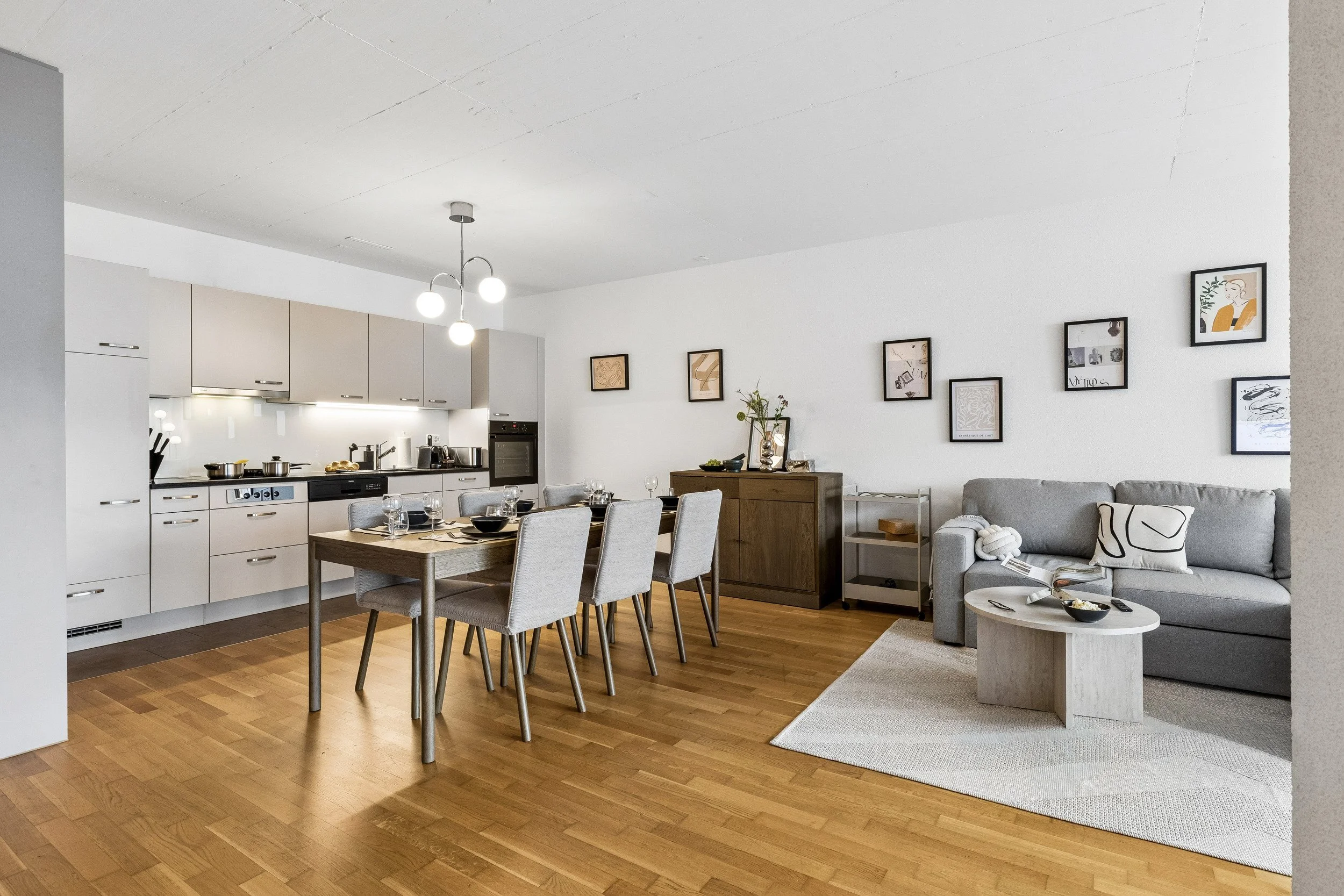 A modern, open-concept living and dining area with white walls, hardwood flooring, a kitchen with gray cabinets and stainless steel appliances, a dining table with six chairs set for a meal, a gray sofa with patterned pillows, a small coffee table wi