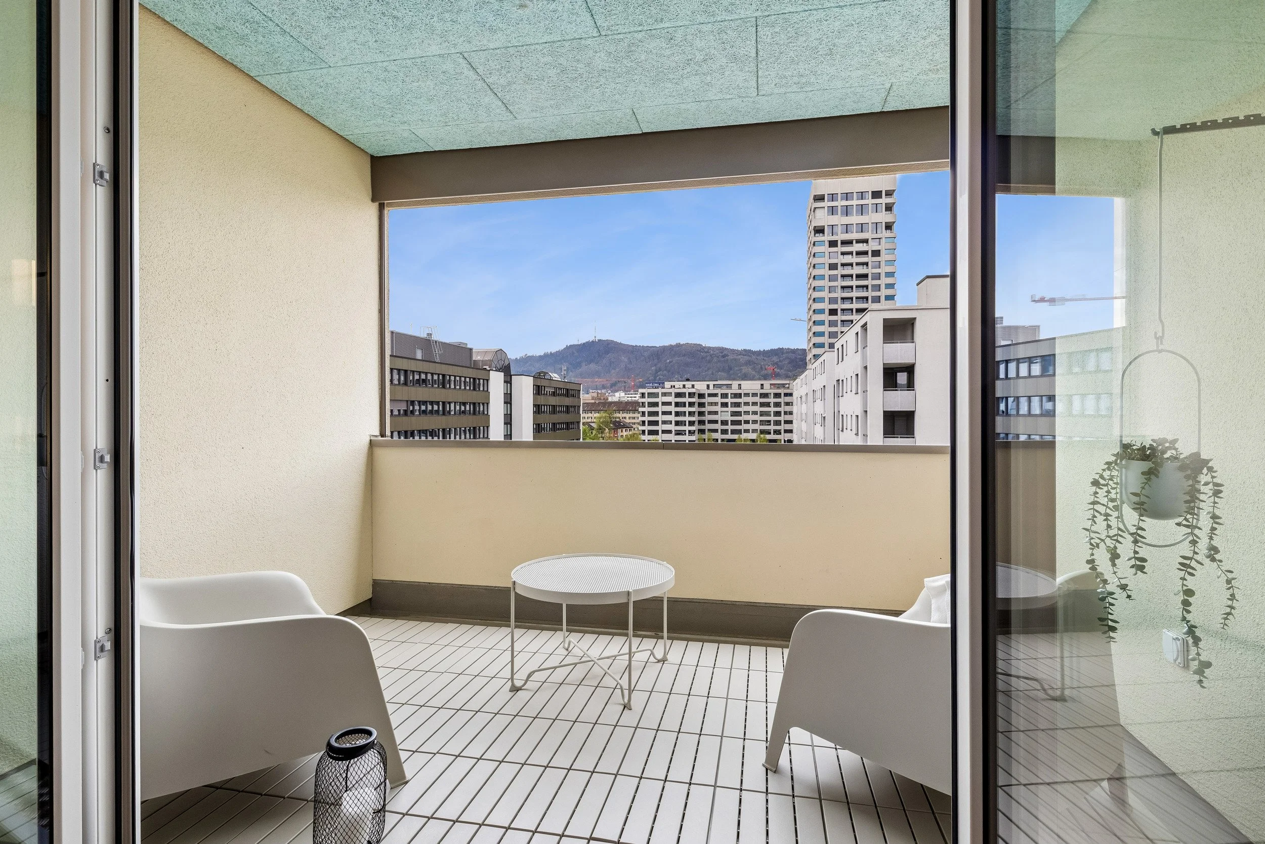 Balcony with white chairs and a small round table, view of city buildings and mountains in the background, seen through glass doors.