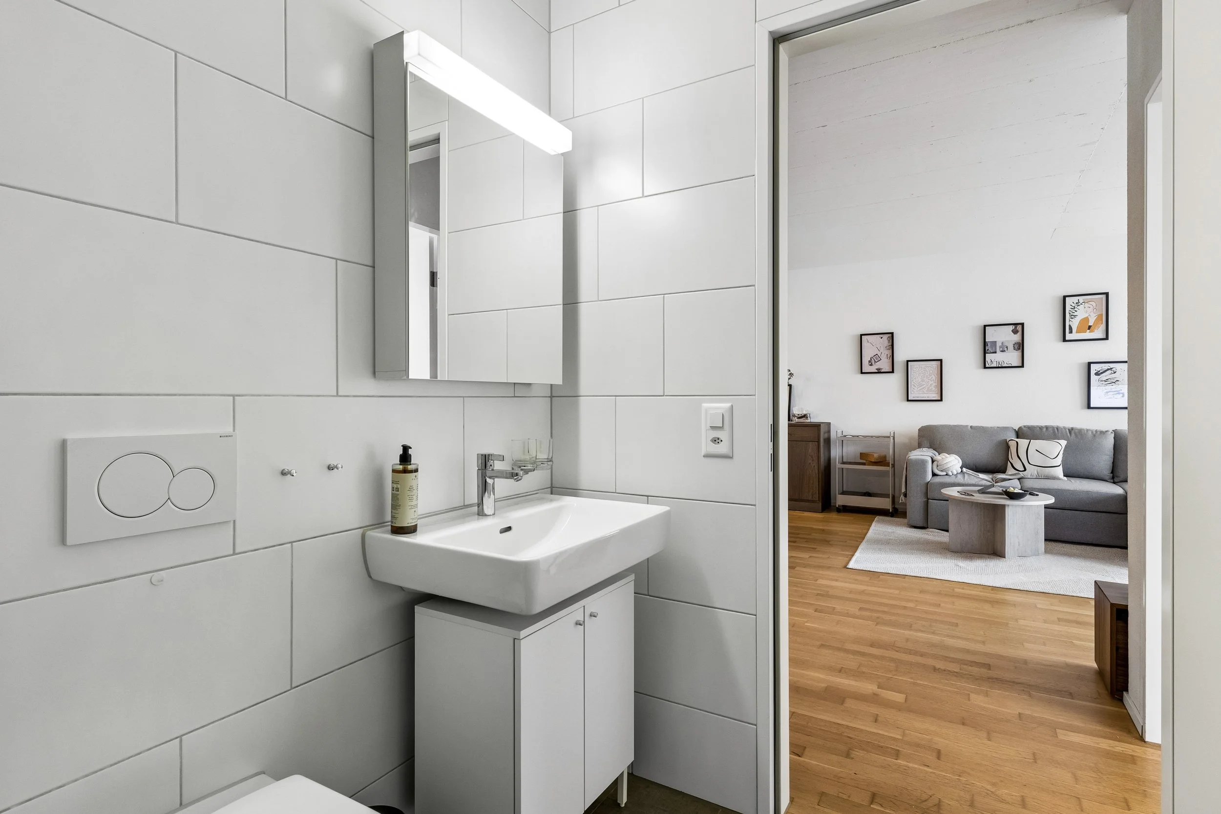 A modern bathroom with white tiled walls, a small sink, and a mirror. The bathroom opens into a living room with wooden floors, a gray sofa, and framed artwork on the wall.