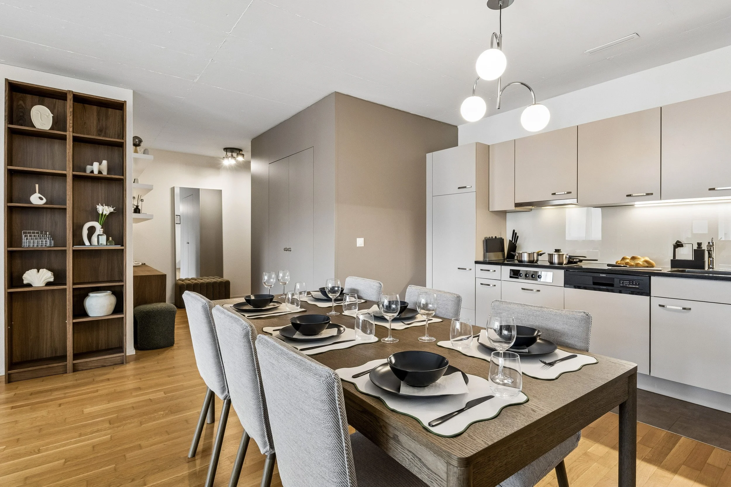 Modern dining area with a wooden table set for dinner with black bowls, glasses, and white placemats, surrounded by light gray upholstered chairs, adjacent to a white kitchen with beige cabinets and stainless steel appliances, and a decorative wooden bookshelf with white vases and ceramics.