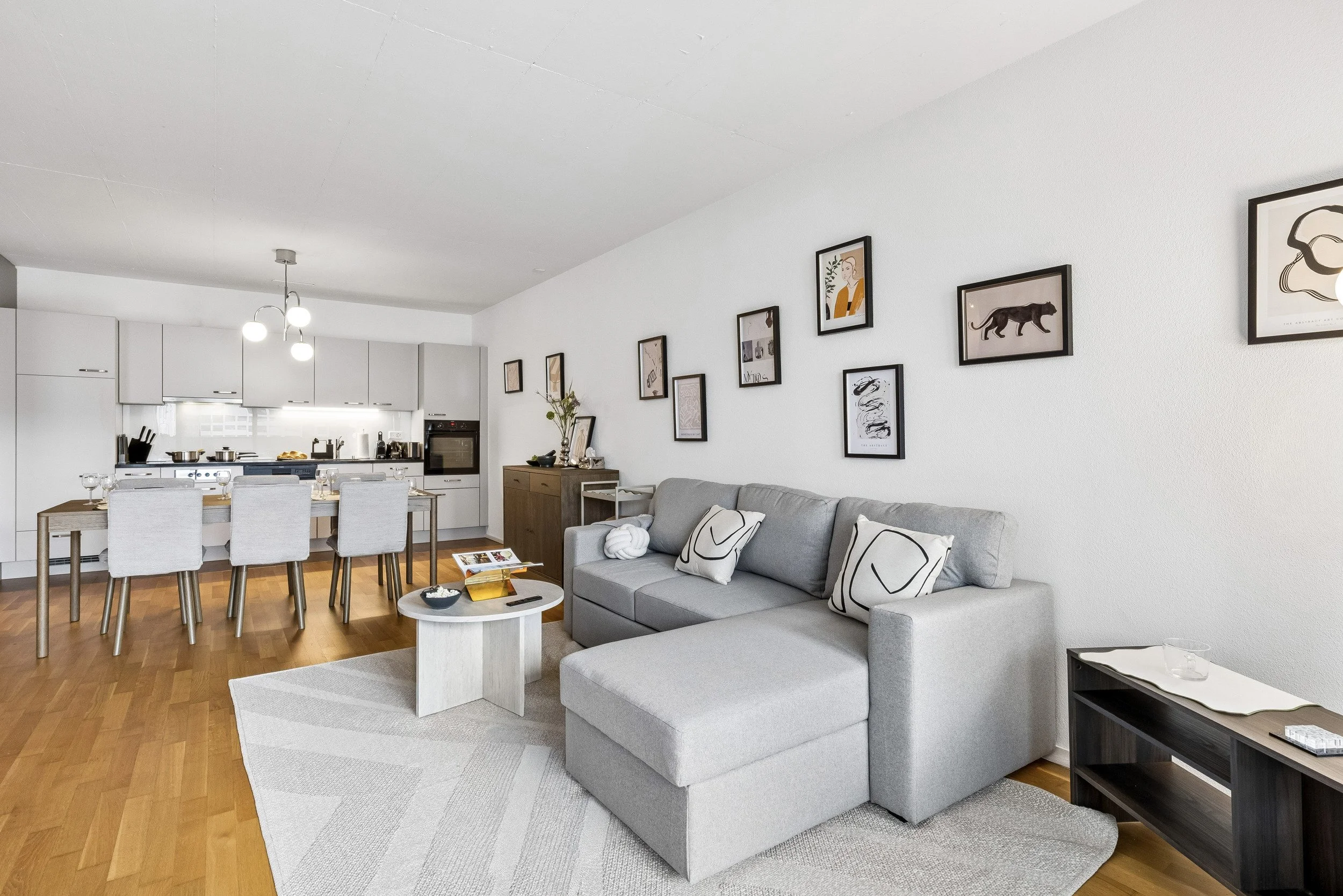 Modern living room with gray sofa, decorative pillows, wooden coffee table, wall art, and open kitchen with dining table and chairs.