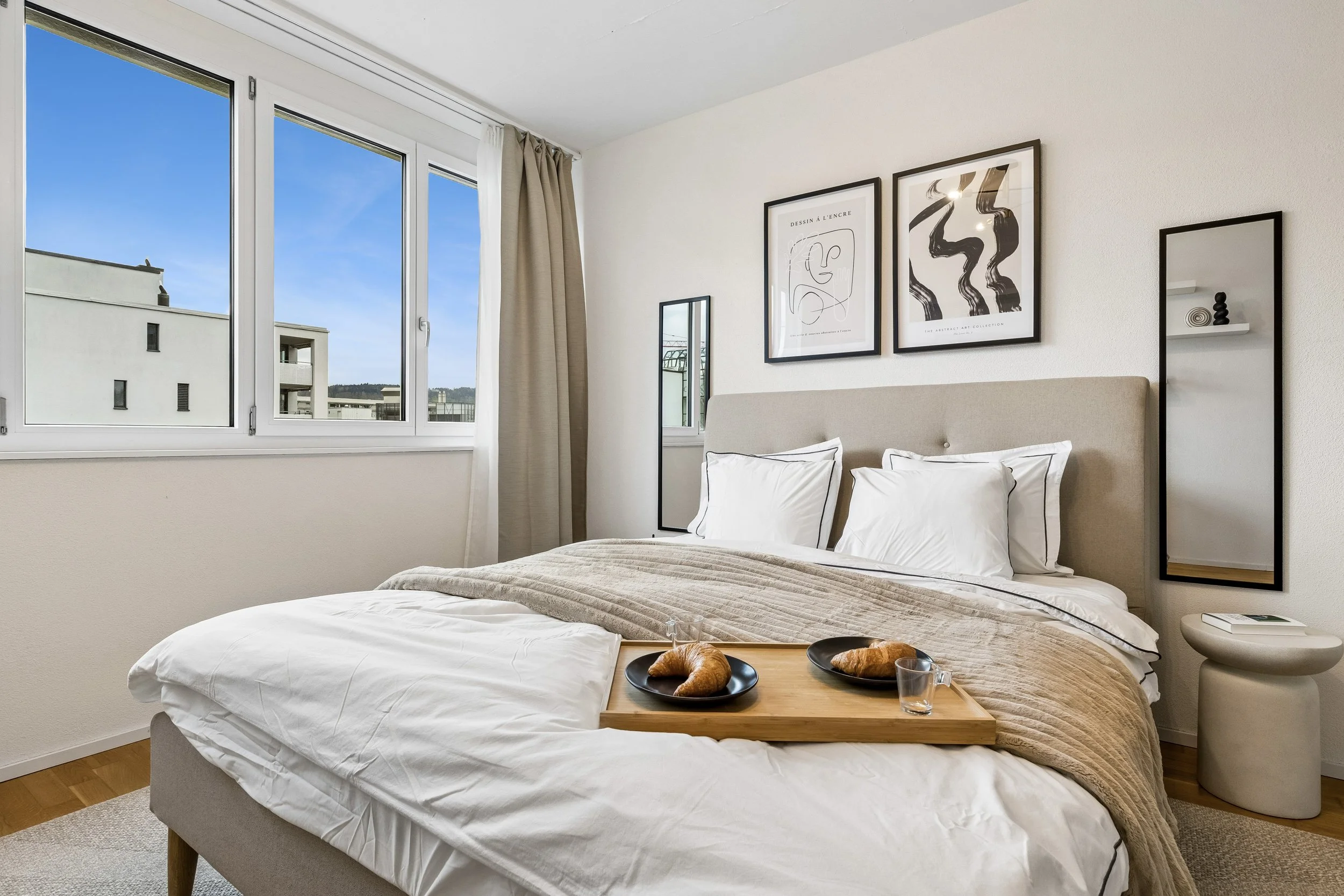 A cozy bedroom with a large window, beige curtains, a bed with a striped beige bedspread, two bedside lamps, and minimalistic wall art.