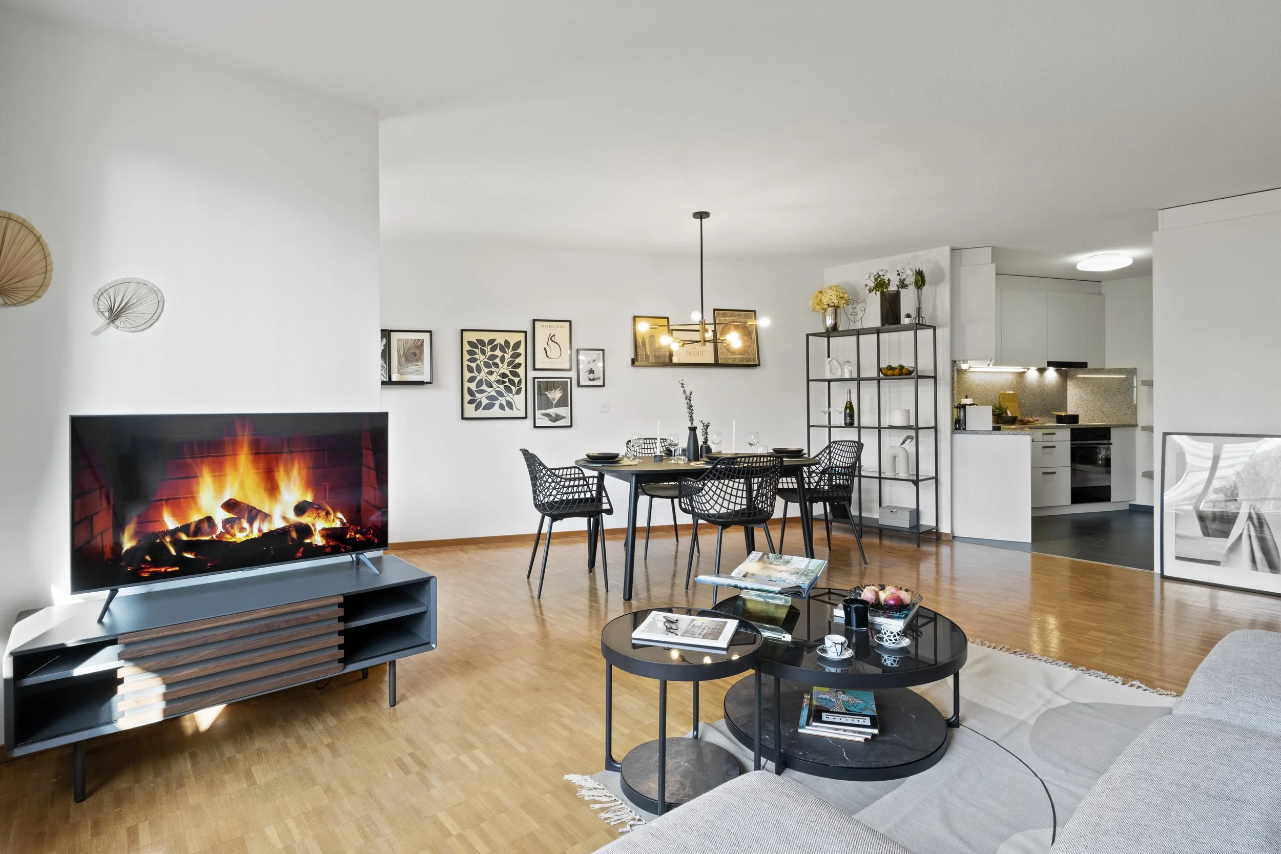 Living room with a fireplace, a black round coffee table with magazines, a white sofa, a dining table with black chairs, wall art, and shelving with plants and decor.