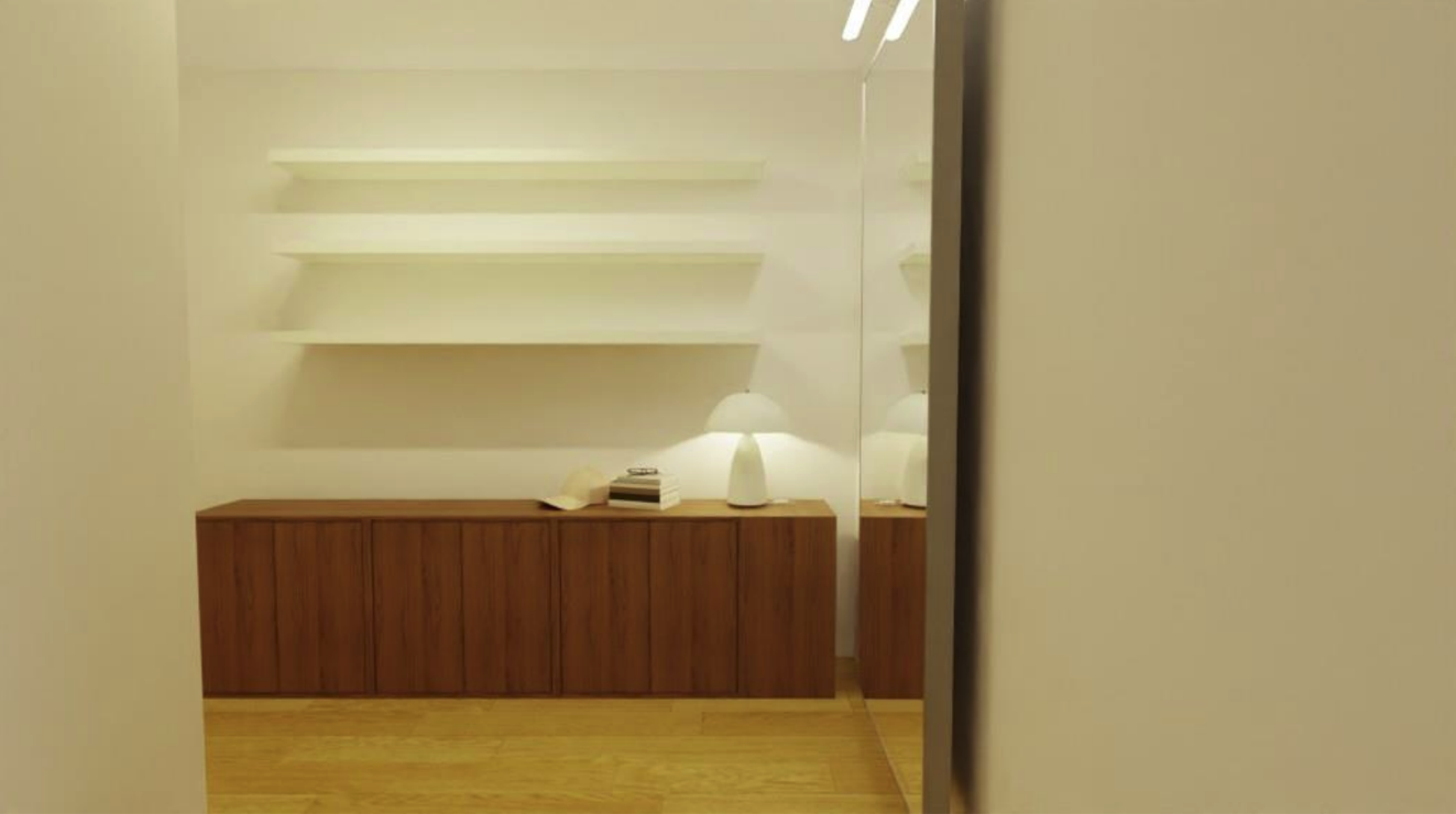 A minimalist indoor space featuring a wooden cabinet with closed doors, a white table lamp, and a few stacked books on top. There are four white floating shelves against the wall above the cabinet. The reflection in a mirror shows part of the room, a