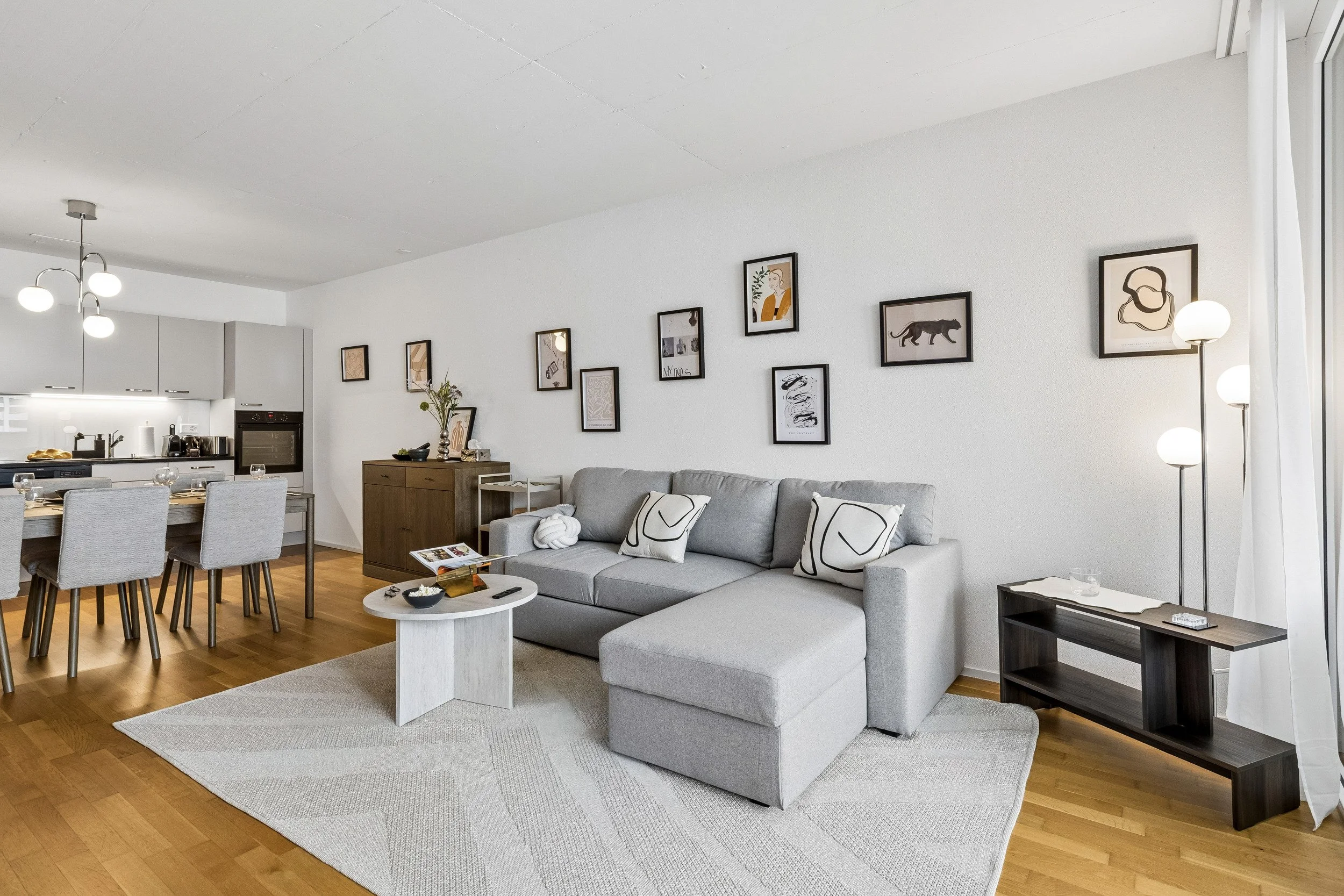 Living room with a gray sofa, framed wall art, a side table, a white coffee table with snacks, and a dining area in the background with a wooden table and gray chairs. The room has wooden flooring, a white textured rug, and a floor lamp with spherica